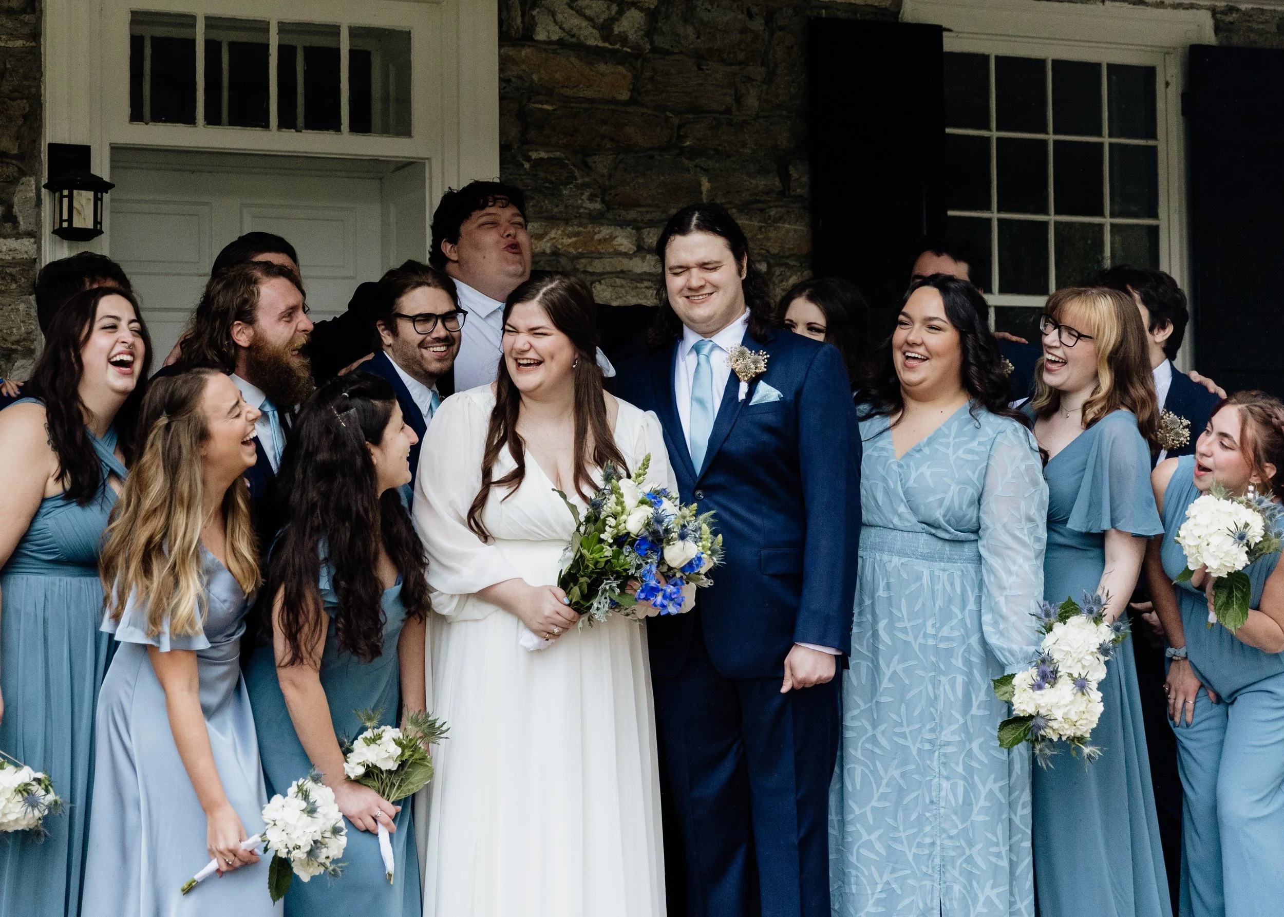 A group of people at a wedding, the bride and groom in the center, surrounded by bridesmaids and groomsmen, all laughing and celebrating outside a stone building.