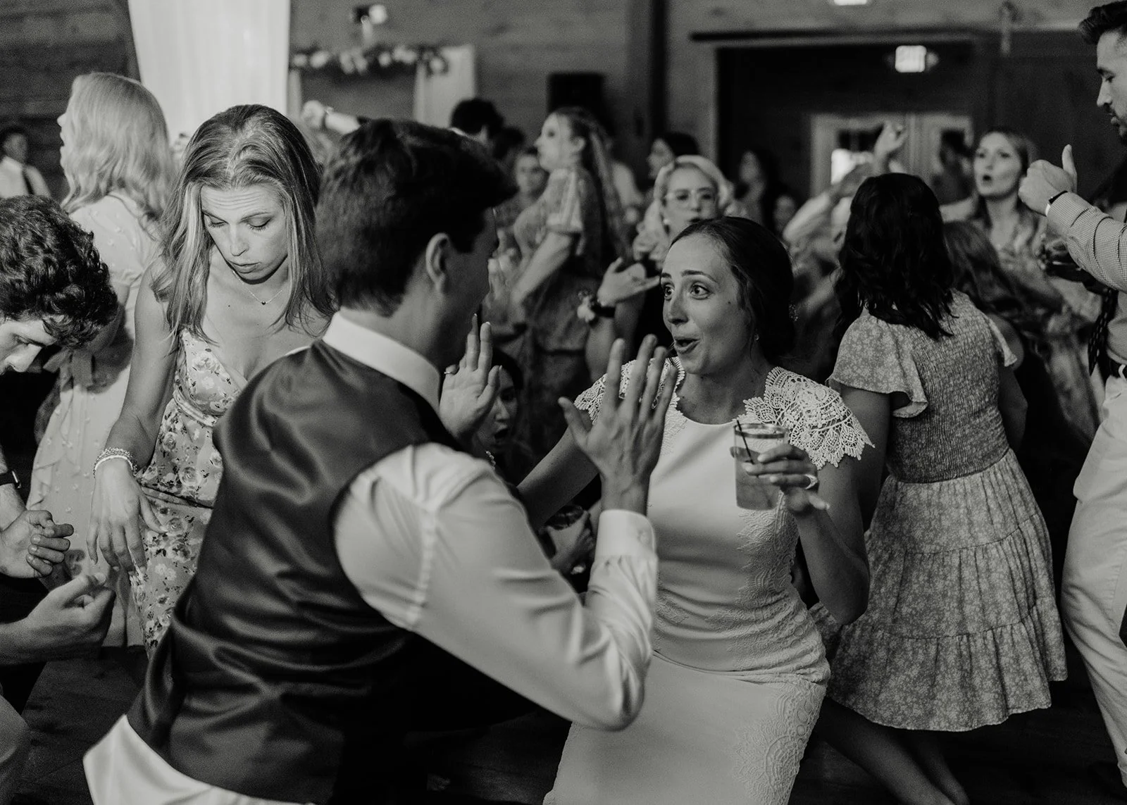 People dancing and socializing at a lively indoor wedding, with some holding drinks, in a rustic venue with wooden walls.