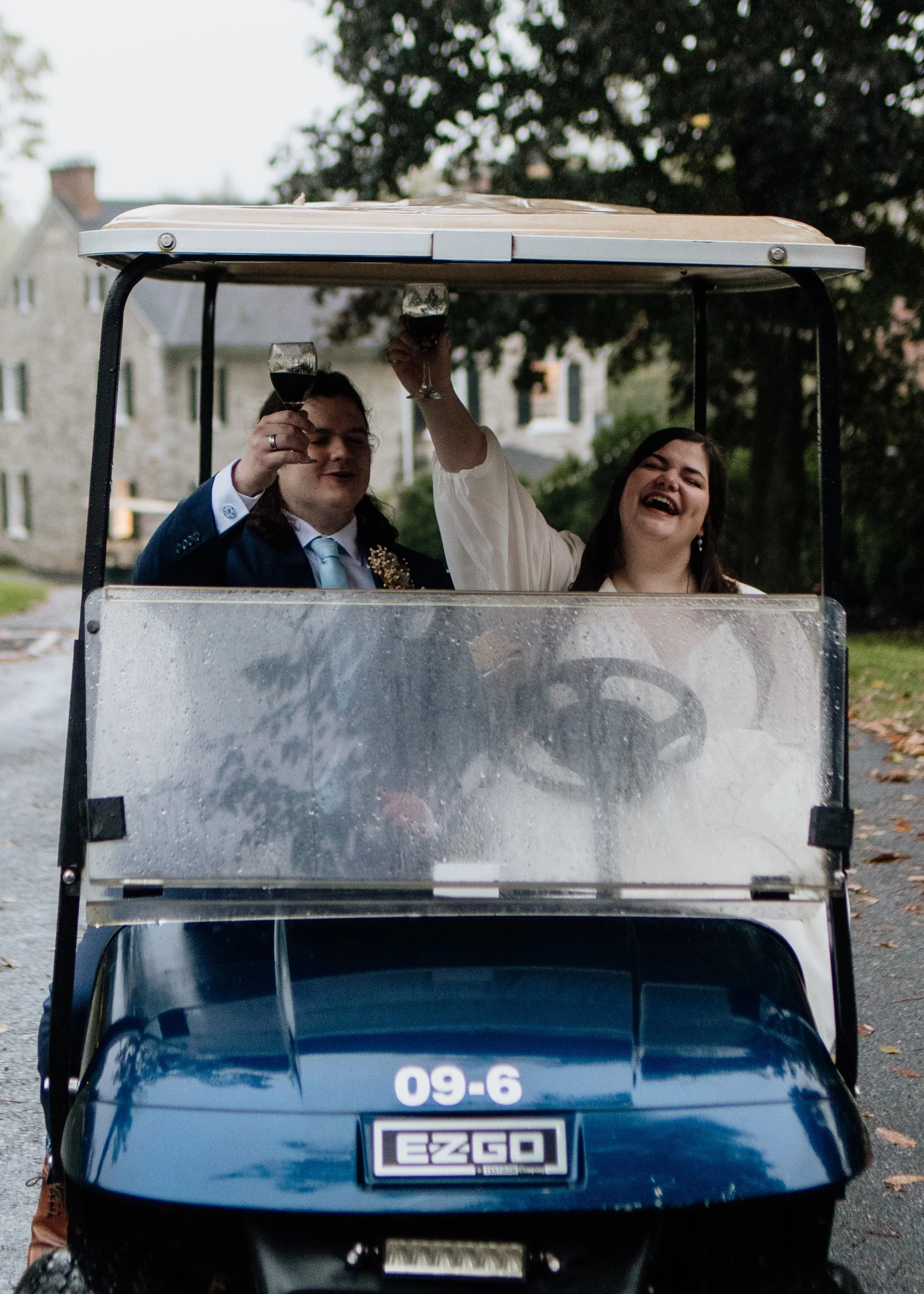 A golf cart with two people seated inside, raising glasses of red wine. The person on the left is dressed in a suit, and the person on the right is laughing. The background shows trees and houses.