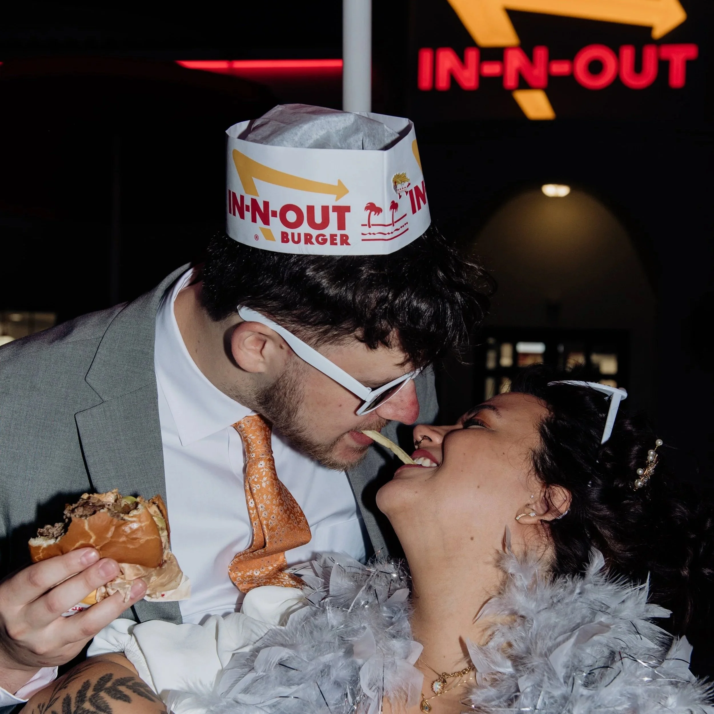 A person holding a half-eaten burger and wearing a paper hat with In-N-Out Burger logo, standing close with another person during a celebration or wedding. The person with the burger is dressed formally in a suit and tie, and the other person is dressed in a white wedding dress with jewelry. The background shows a neon In-N-Out sign.