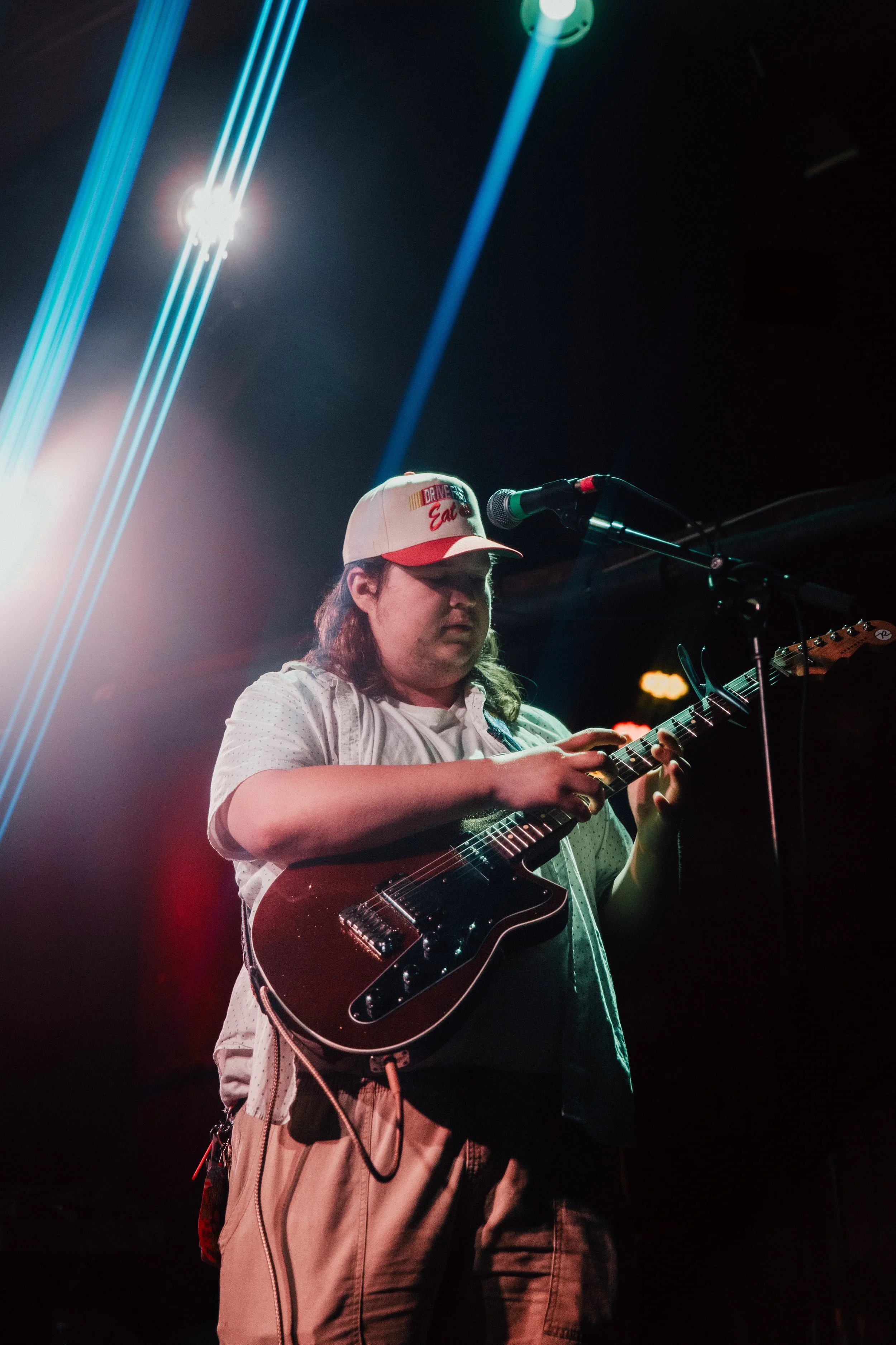 A person playing an electric guitar on stage, wearing a white cap, a light-colored shirt, and beige pants, with microphone on stage and dramatic stage lighting effects.