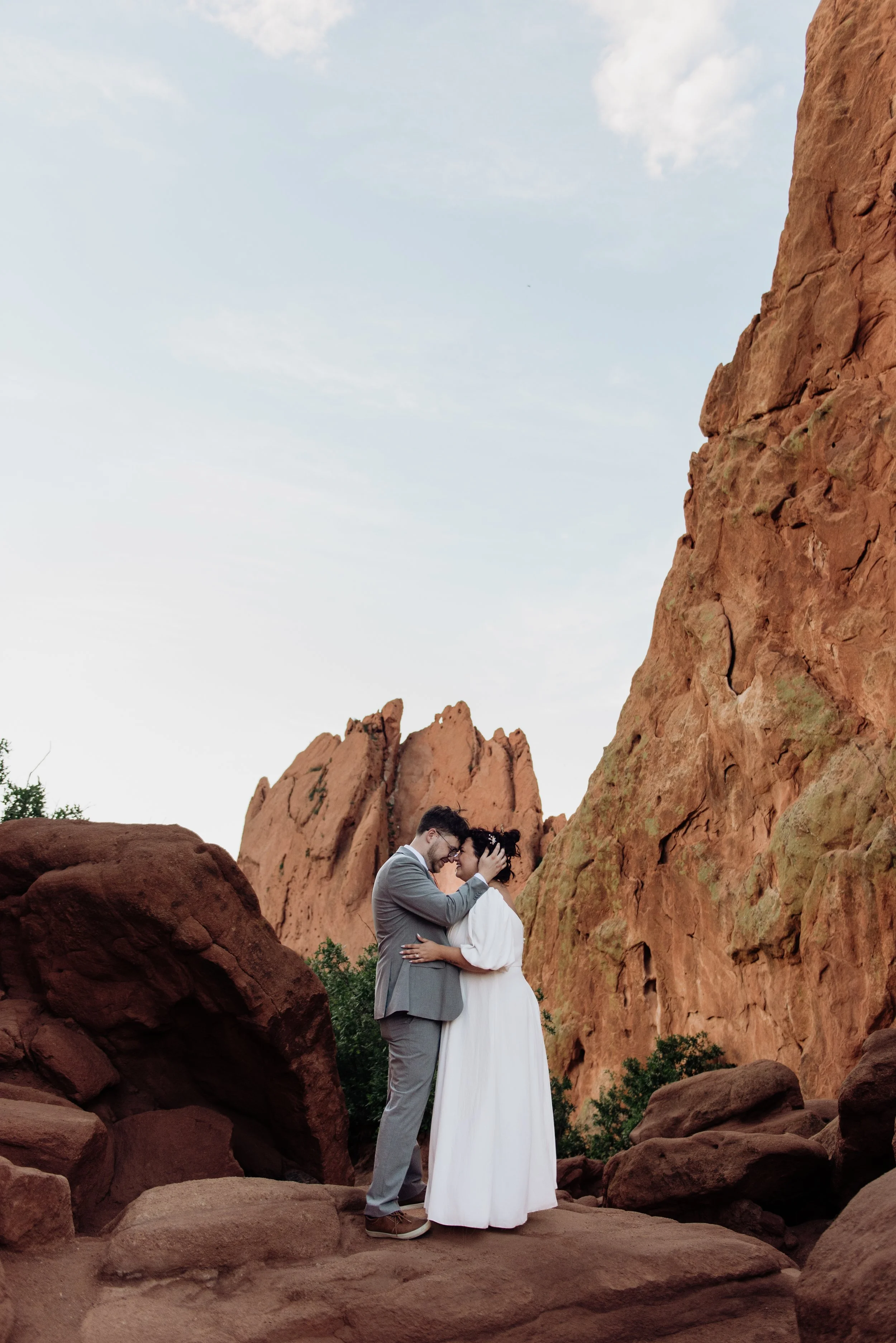 A couple dressed in wedding attire, standing close and touching foreheads in a rocky outdoor setting with red cliffs and a partly cloudy sky.