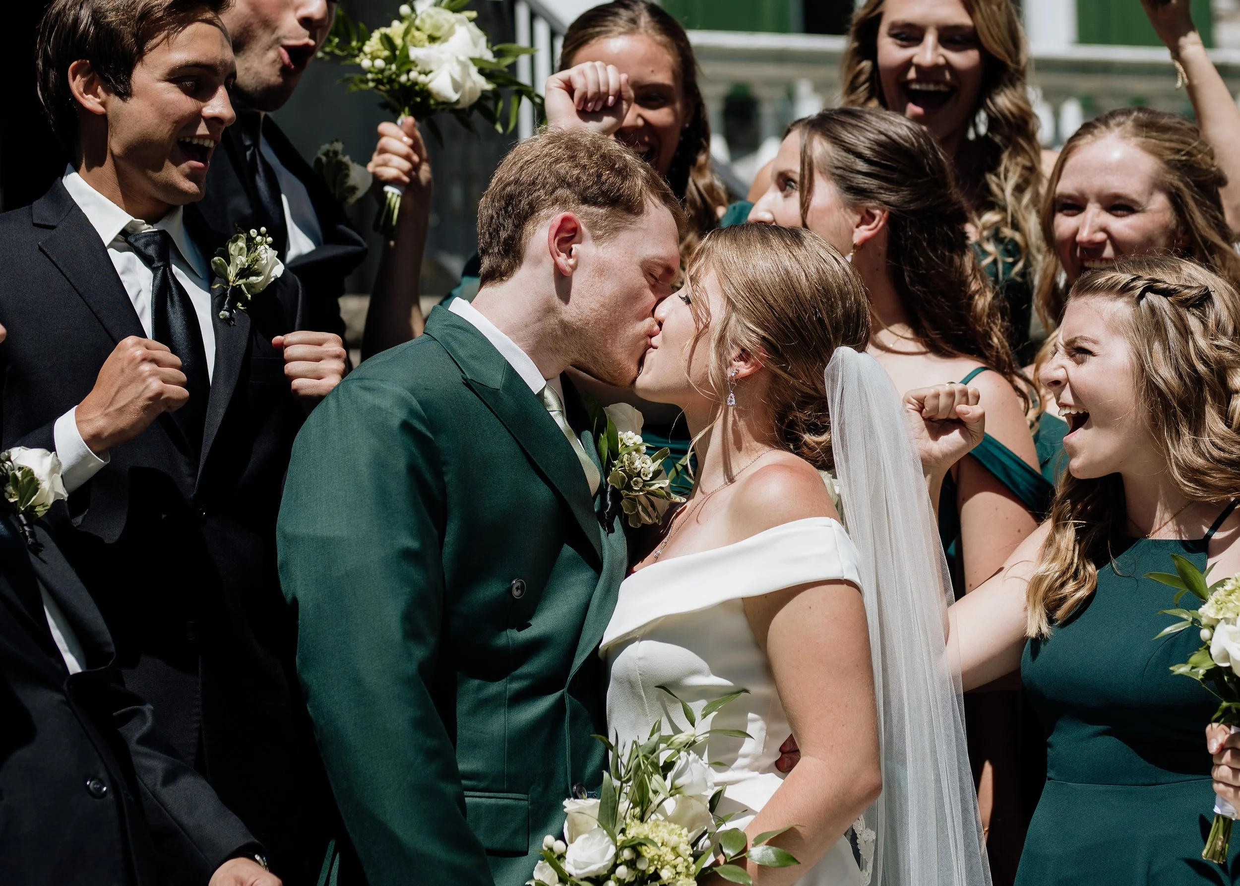 A newlywed couple kissing during their wedding ceremony surrounded by happy friends and family, outdoors on a sunny day.