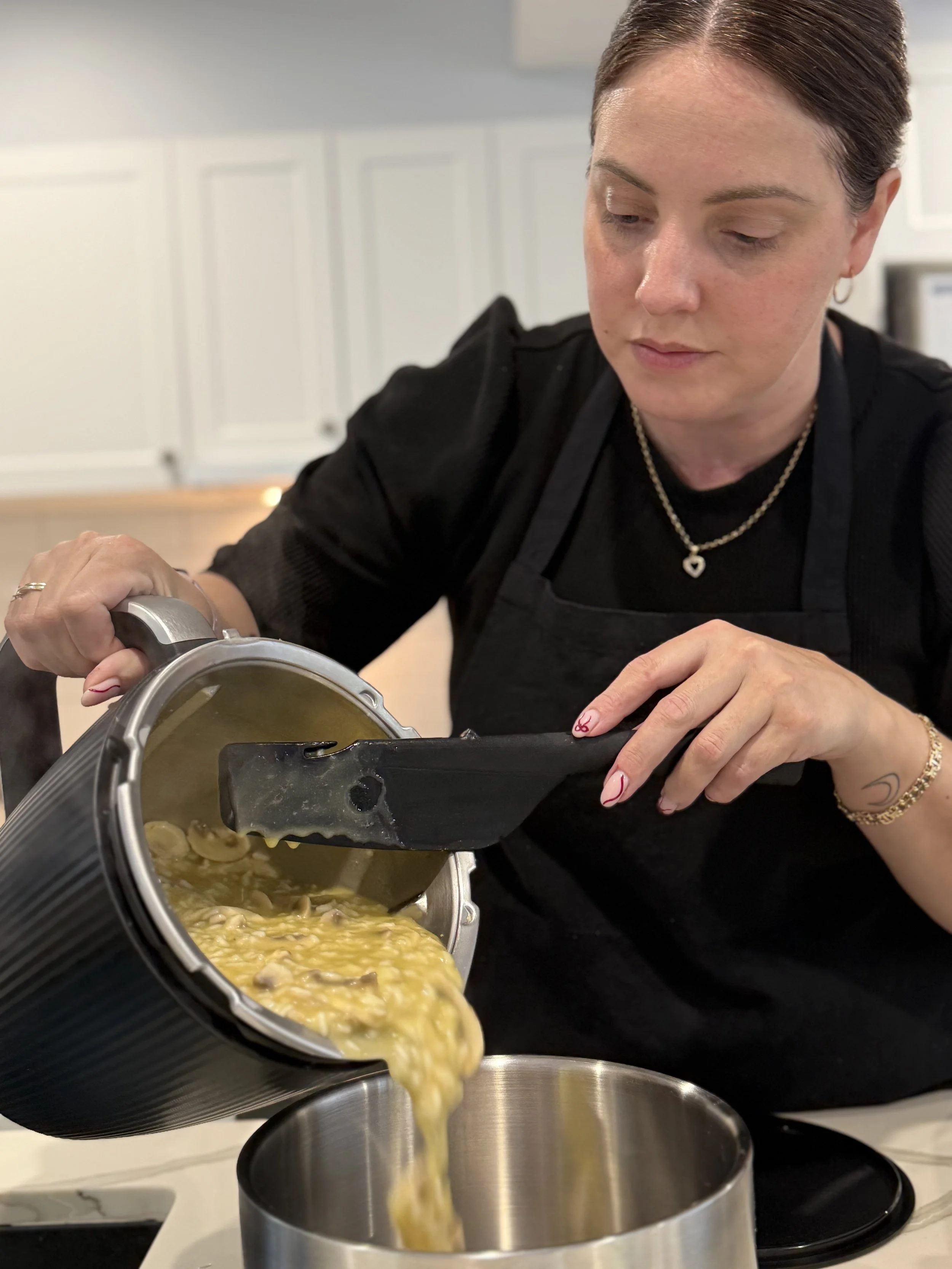 A woman with short brown hair, wearing a black top, jewelry, and earrings, is pouring a can of soup into a pot in a kitchen.