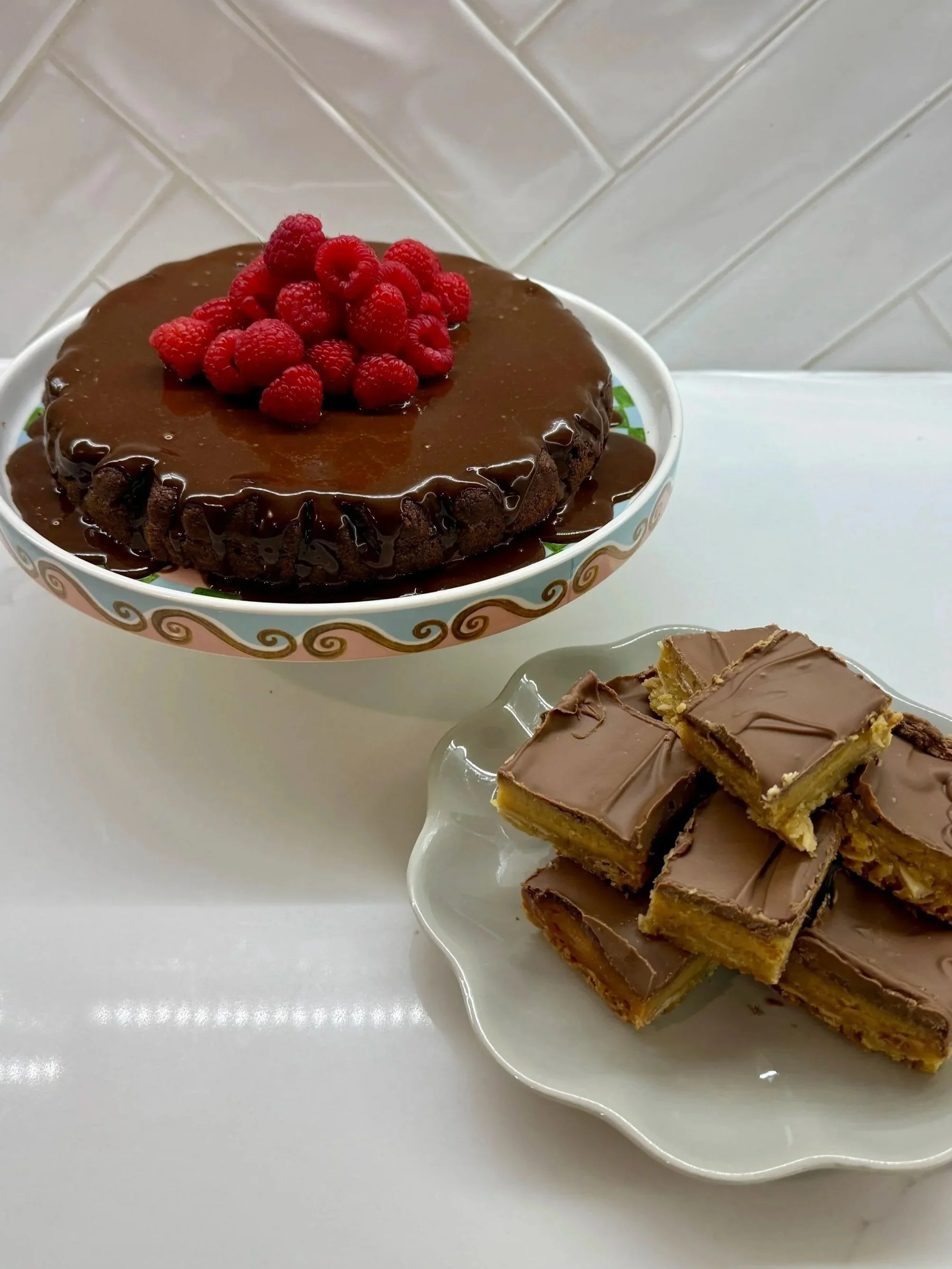 A chocolate cake topped with raspberries on a decorative plate, with a plate of layered chocolate-coated bars nearby on a white table against a tiled wall background.