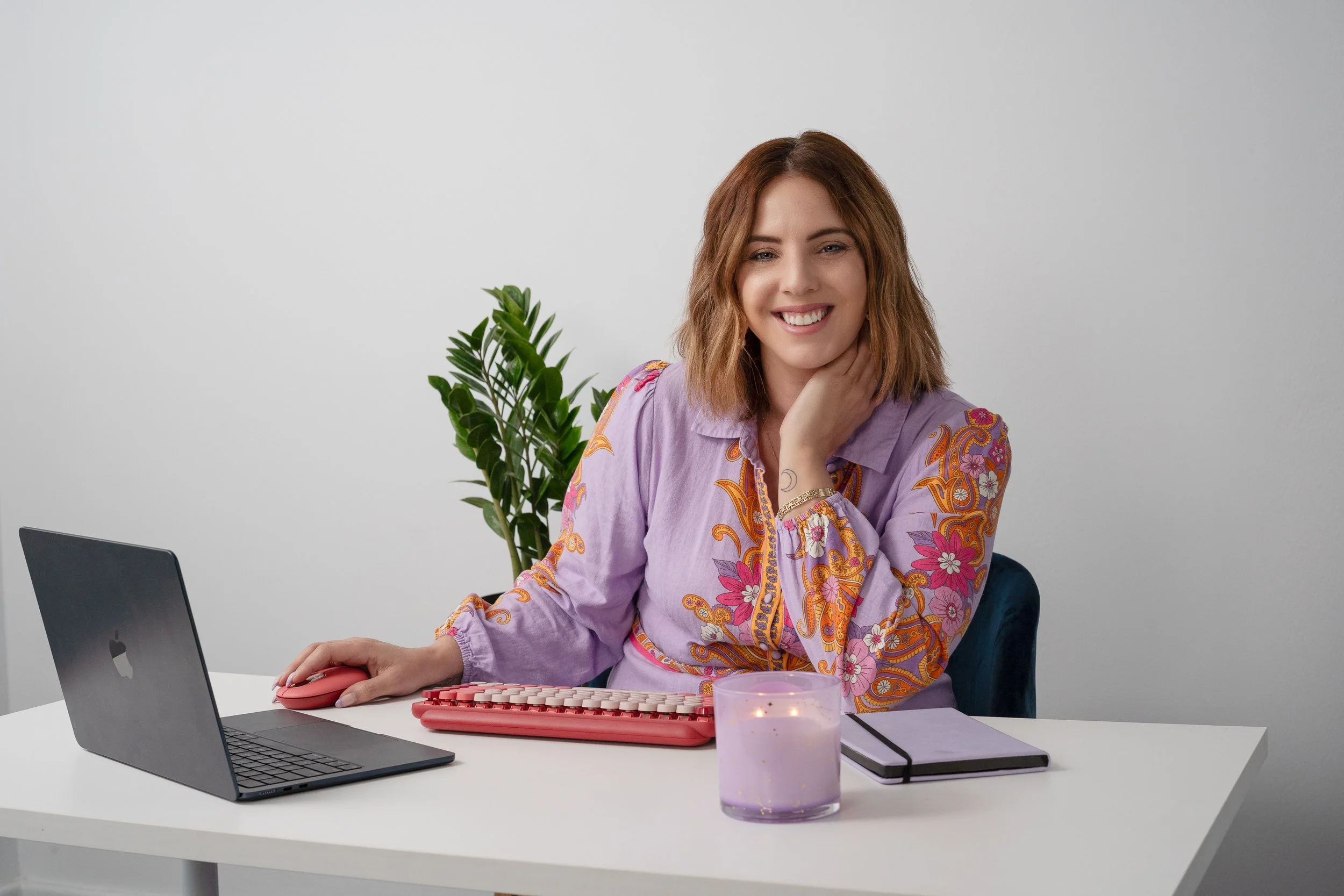 A woman with shoulder-length brown hair sitting at a white desk, smiling, with a gray wall behind her. She is wearing a purple shirt with colorful floral embroidery, holding a pink computer mouse. On the desk, there is a partially open laptop, a purple candle, some papers, and a closed notebook. There is a leafy green plant behind her.