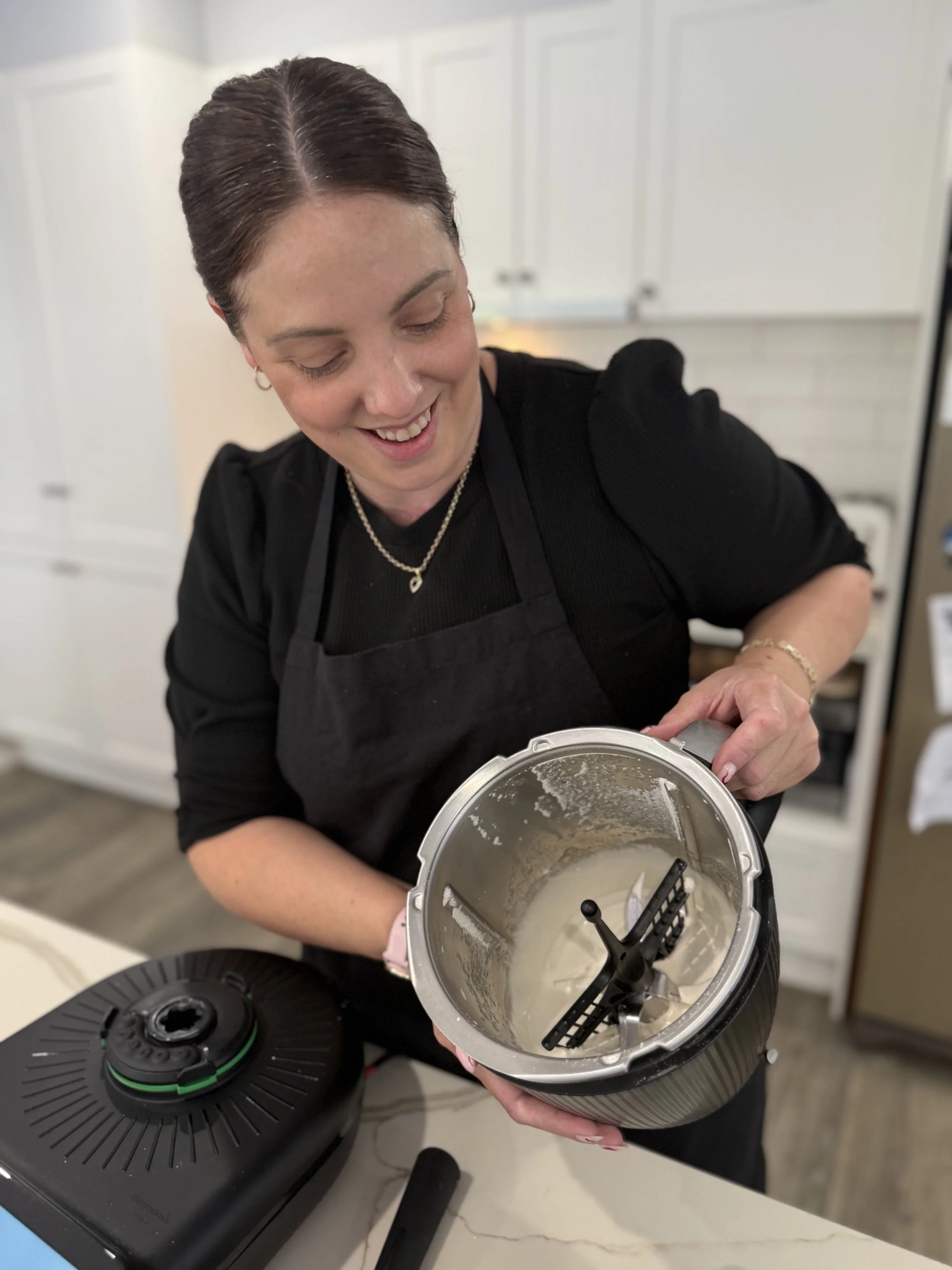 A woman in a black shirt and apron is smiling and looking down while holding a cooking appliance with a mixture inside. There is a black kitchen appliance on the countertop in front of her.