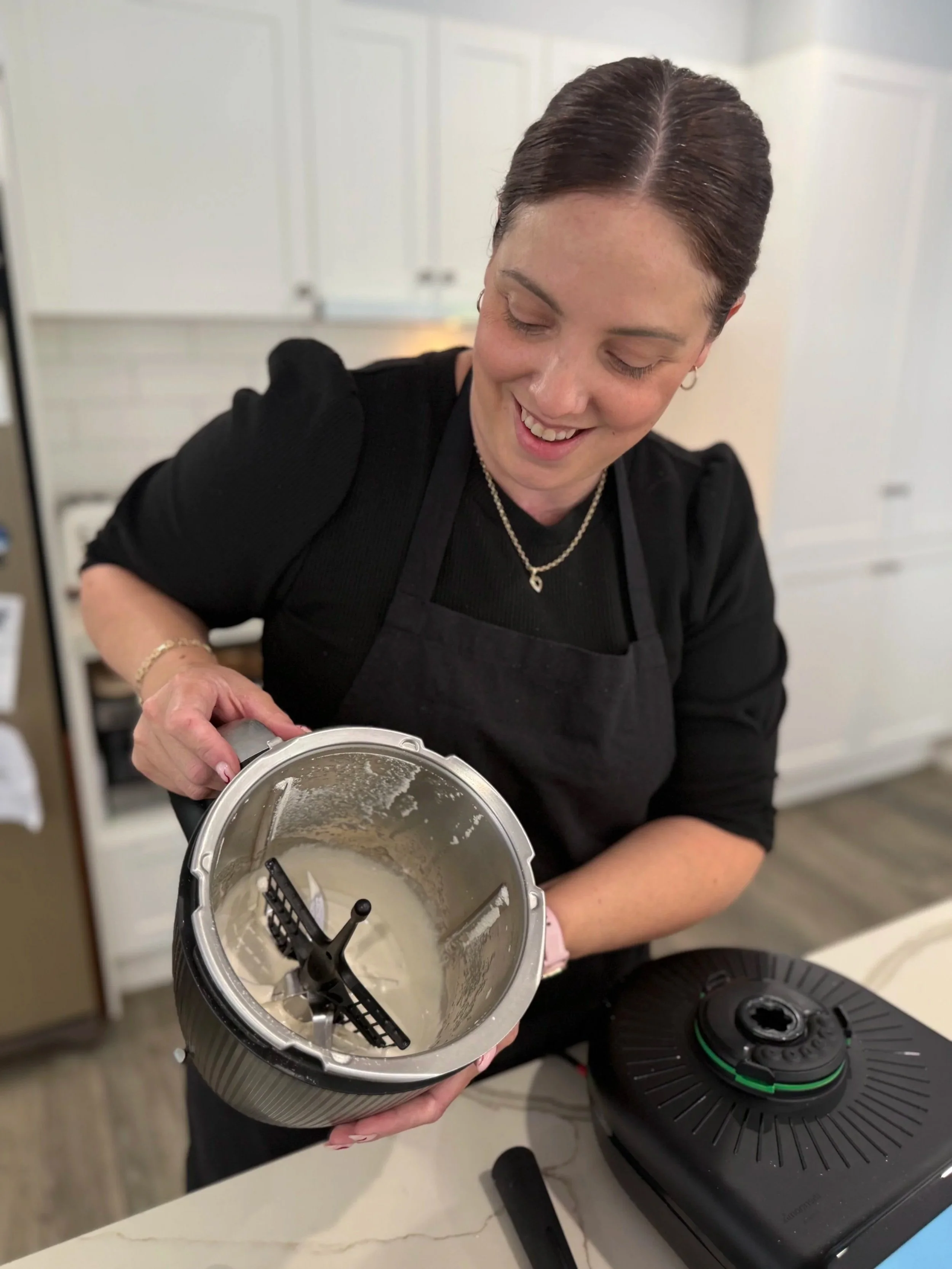 A woman in a black shirt and apron is smiling while holding the lid of a blender filled with a creamy mixture. She is in a kitchen with white cabinets in the background and an electric stove on the counter.