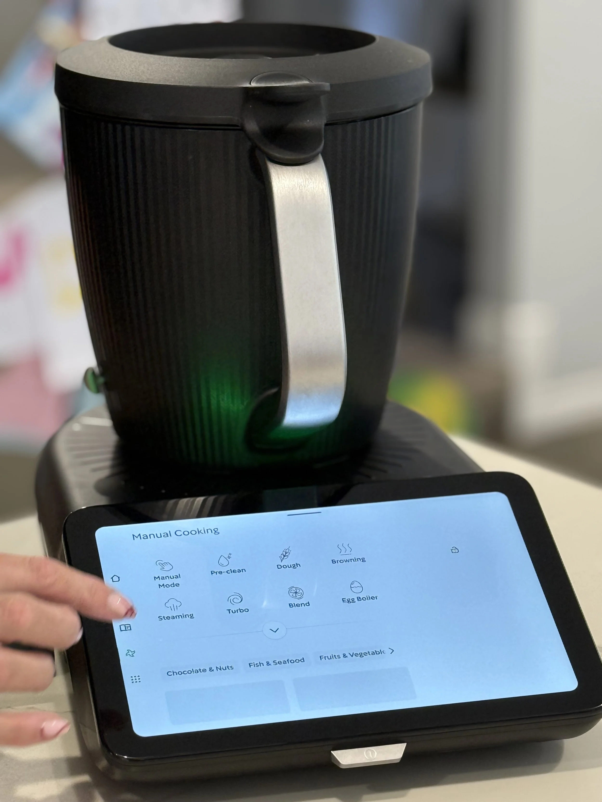 A Thermomix machine with a digital touchscreen interface displaying manual cooking options, with a person's hand pointing at the screen.