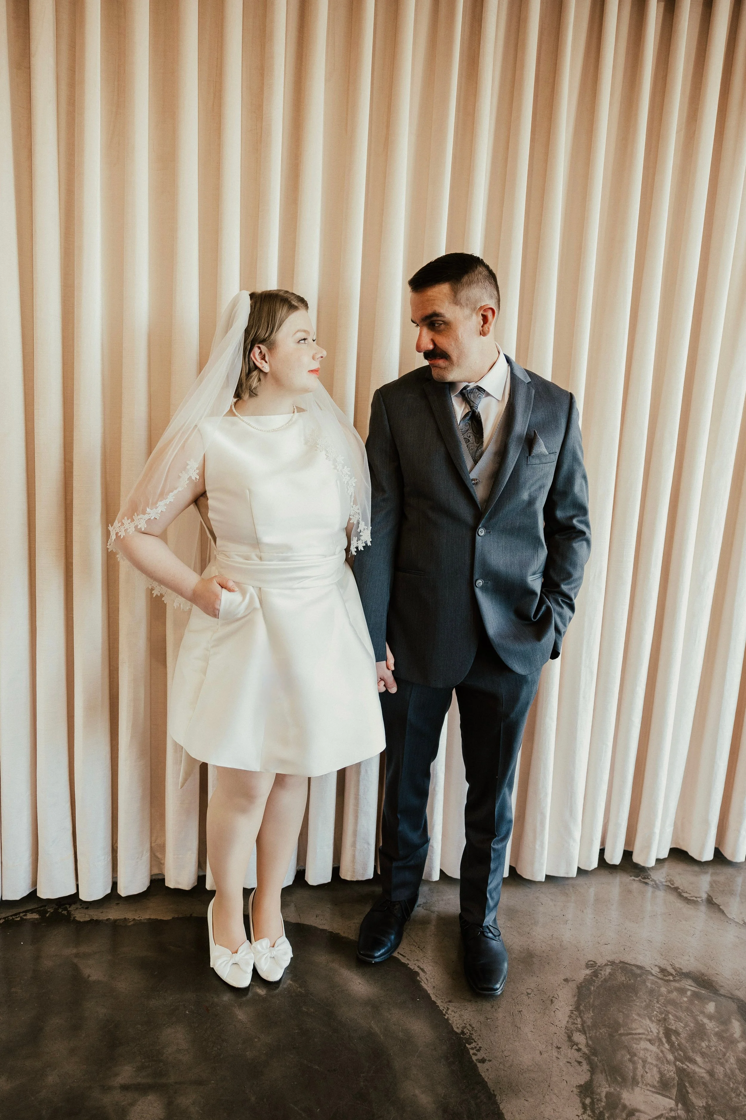 Bride and groom standing together, holding hands, facing each other, in front of a beige curtain.