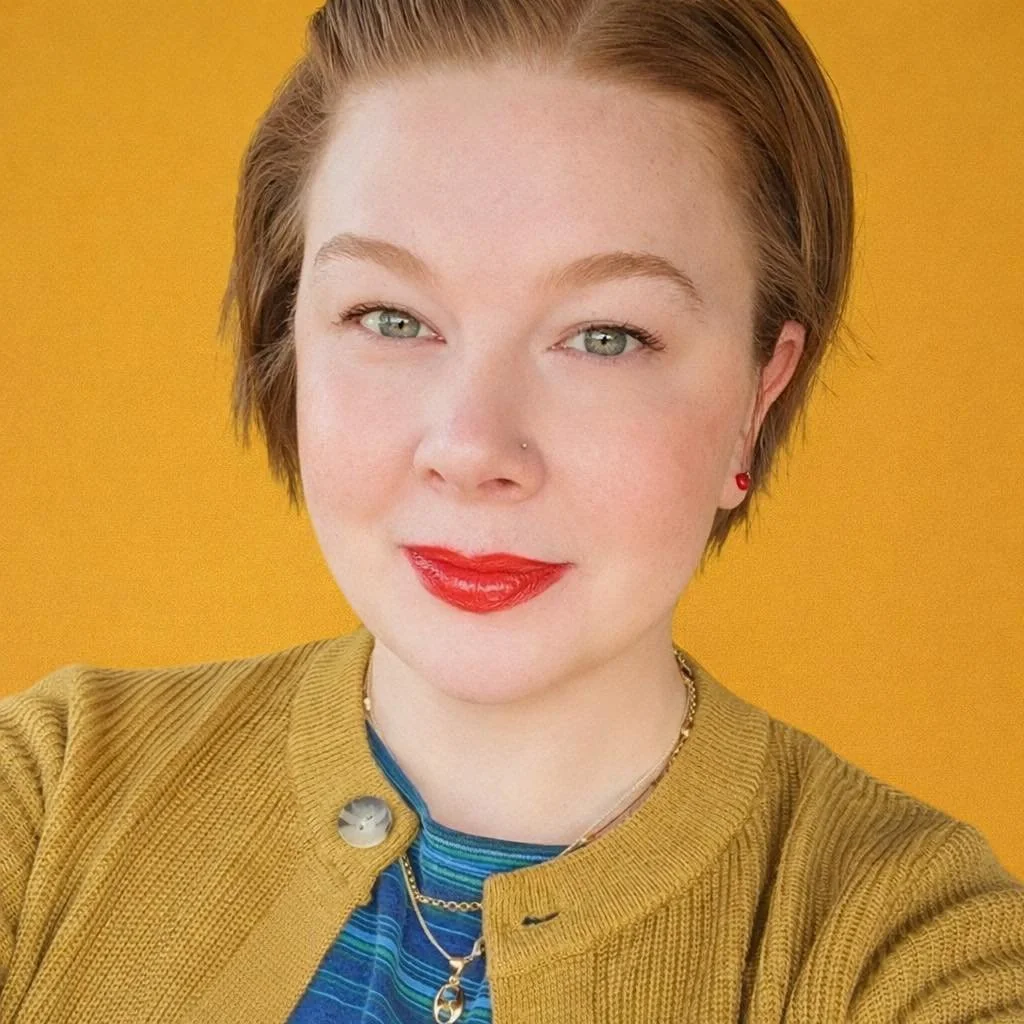A young woman with short, reddish-brown hair and fair skin, wearing a yellow cardigan over a blue striped shirt, red earrings, and red lipstick, standing against a yellow background.