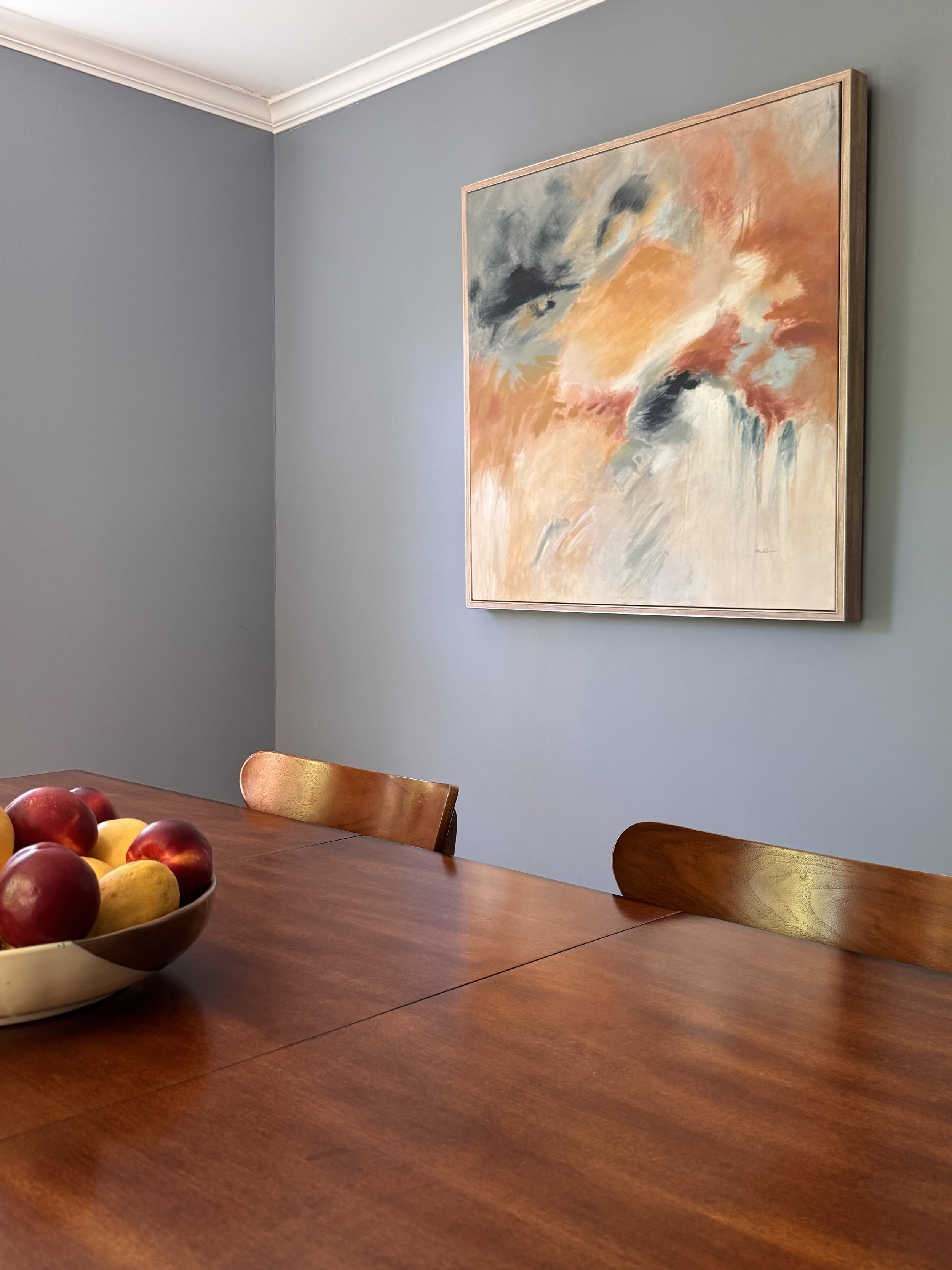 A wooden dining table with a bowl of apples and peaches, surrounded by wooden chairs, with a blue wall, crown molding, and an abstract painting hanging on the wall.
