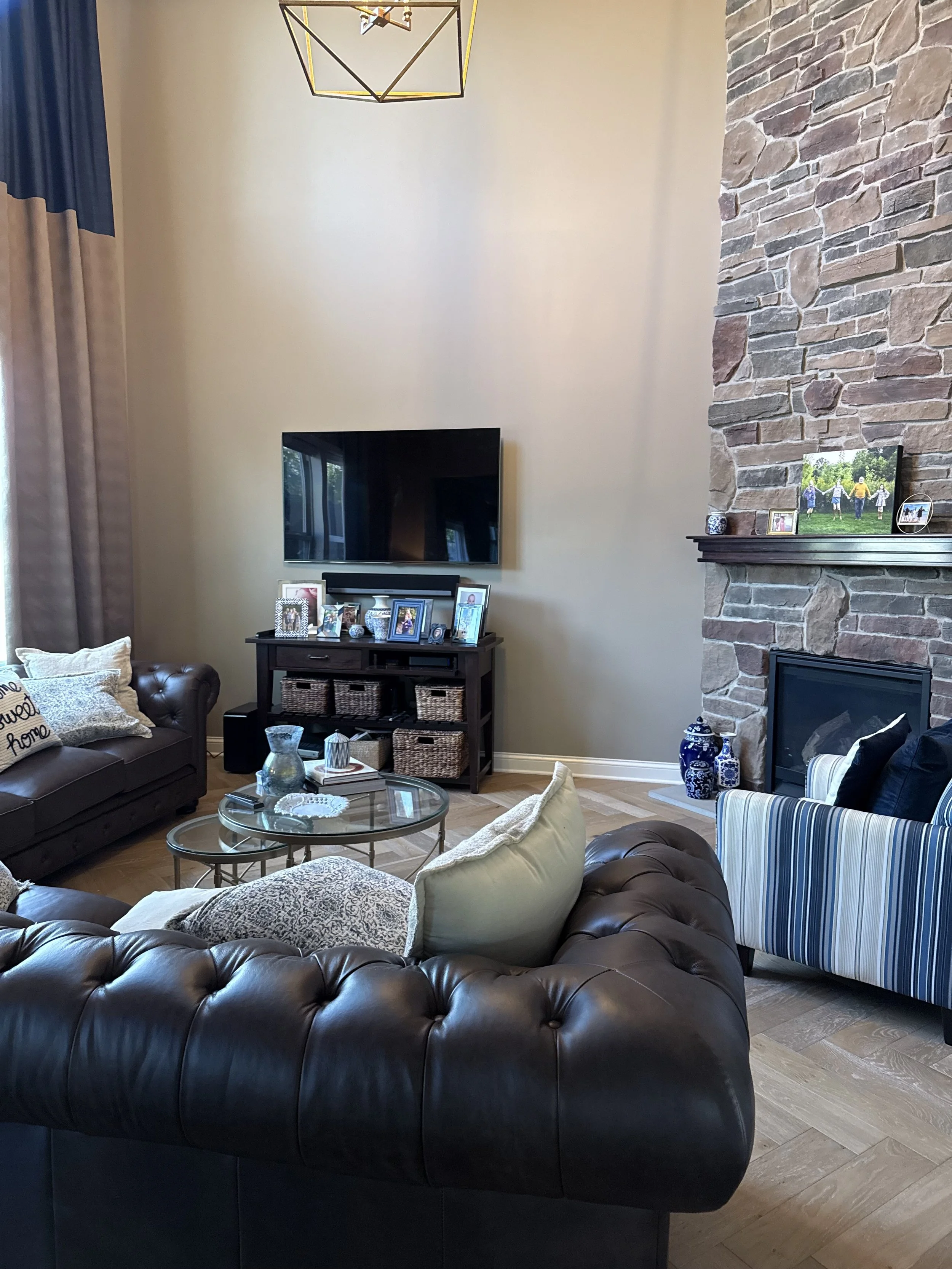 Living room with leather sectional sofa, striped accent chair, glass coffee table, wall-mounted TV, fireplace with brick surround, framed photos and decorative items on mantel, beige wall, and dark hardwood flooring.