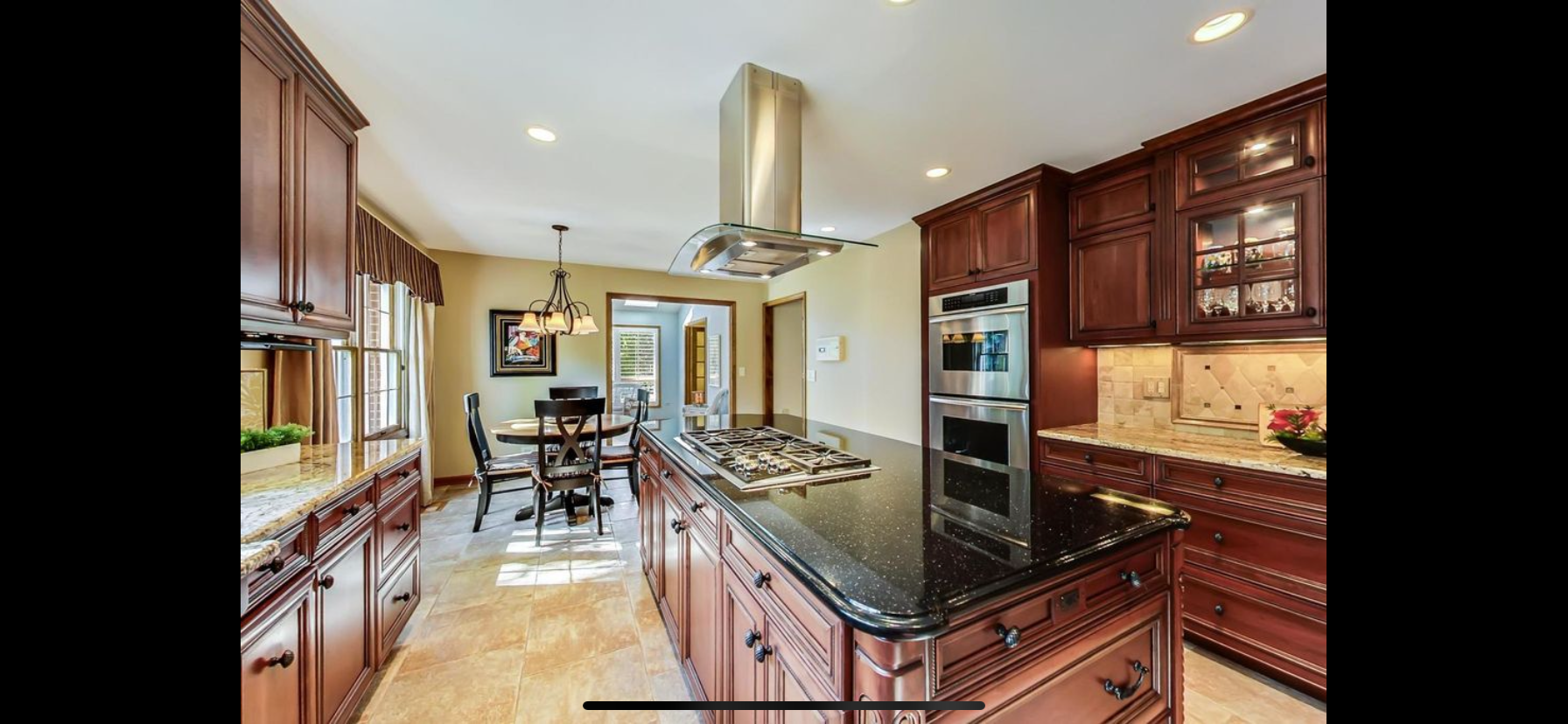 A spacious kitchen with dark wood cabinetry, a black granite countertop island with a gas stovetop, stainless steel double ovens, and a dining area with a table, chairs, and a chandelier in the background.