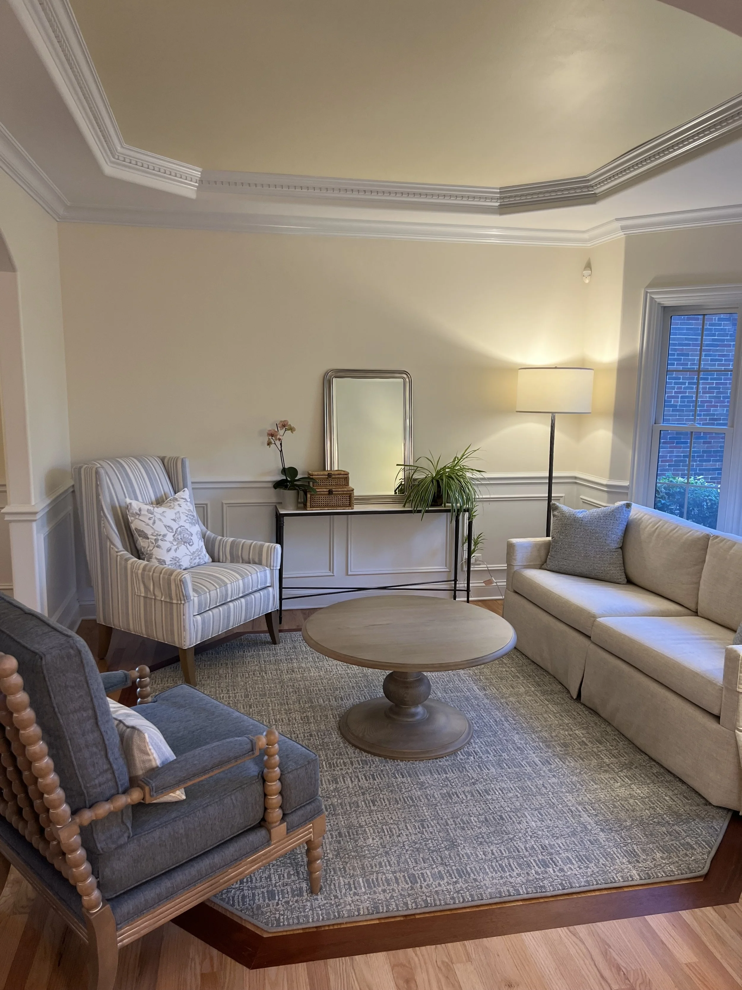 Living room with beige walls and ceiling, white crown molding, a beige sofa with gray pillows, a gray armchair with floral patterned cushions, a wooden coffee table, a black console table with a mirror, plants, and a floor lamp near a window with blue brick wall outside.