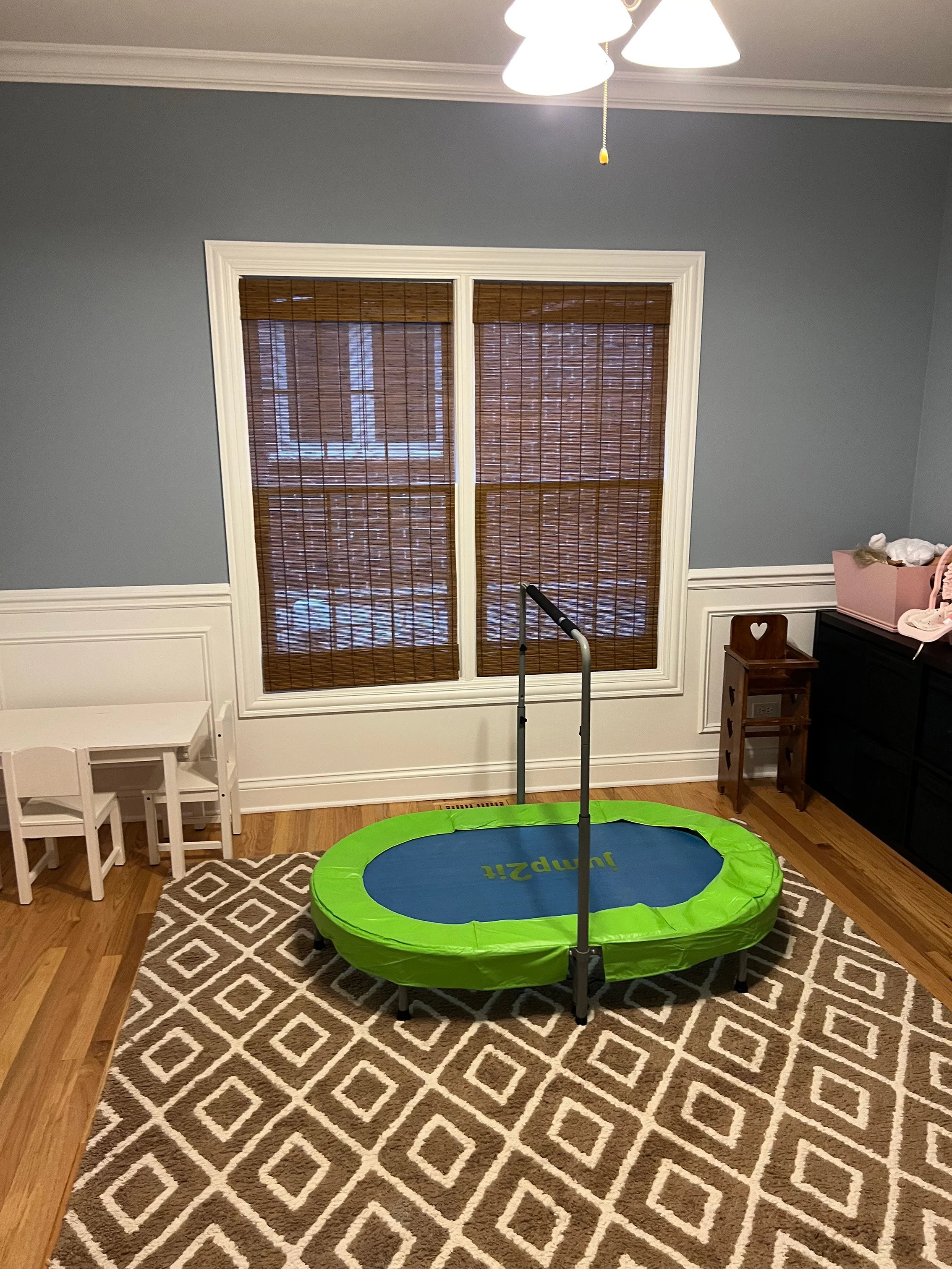 A room with brown bamboo window blinds, a patterned brown and white rug, a small white children’s table with two chairs, a black dresser with a pink container on top, a small wooden shelf with heart cutouts, a part of a trampoline with a black pole, and a chandelier with three white lamps held by a yellow pull cord.