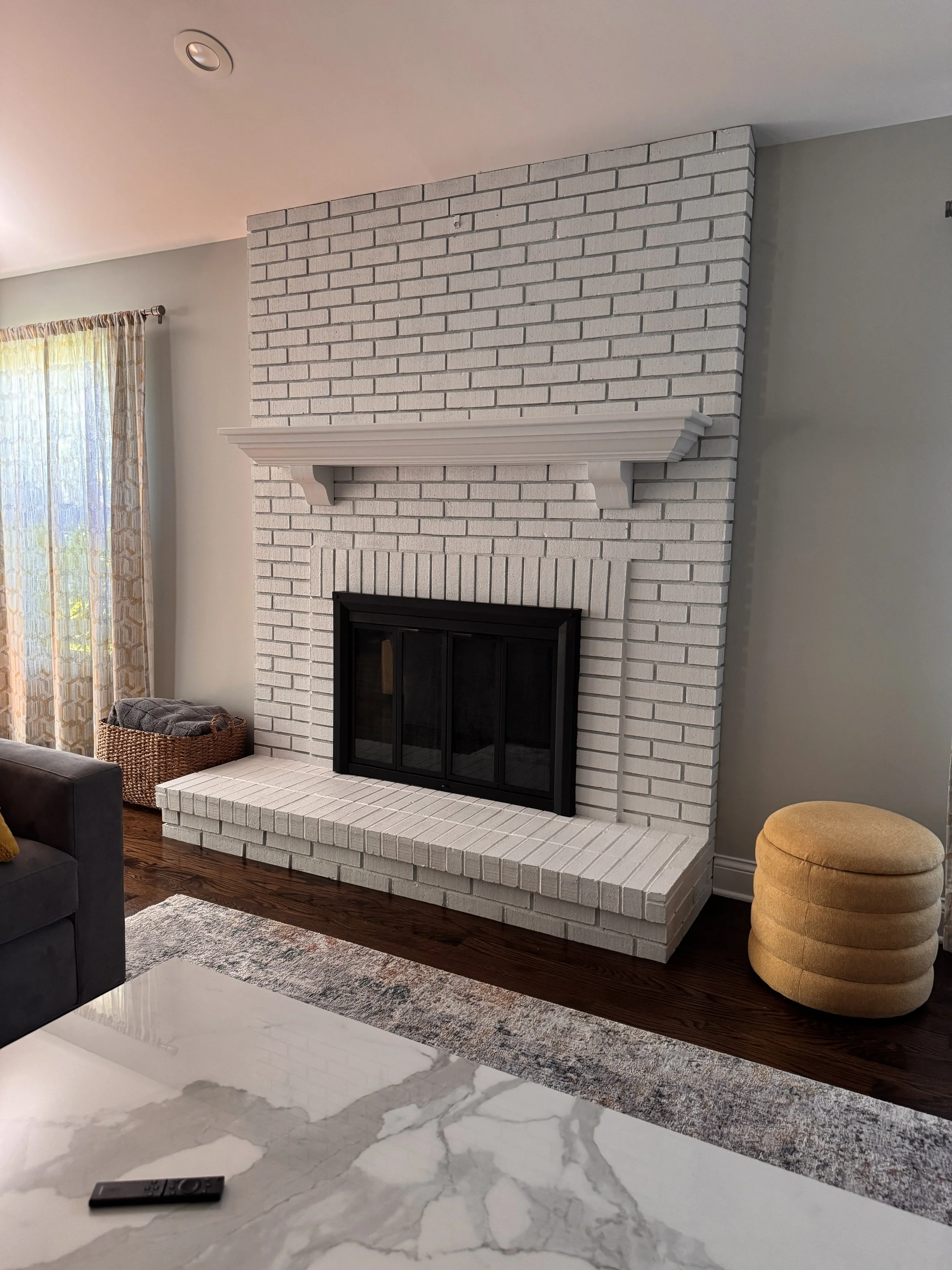 Close-up of a white brick fireplace with a mantel, in a living room with a gray wall, yellow ottoman, and a partial view of a dark gray sofa, marble floor, and remote control on the floor.