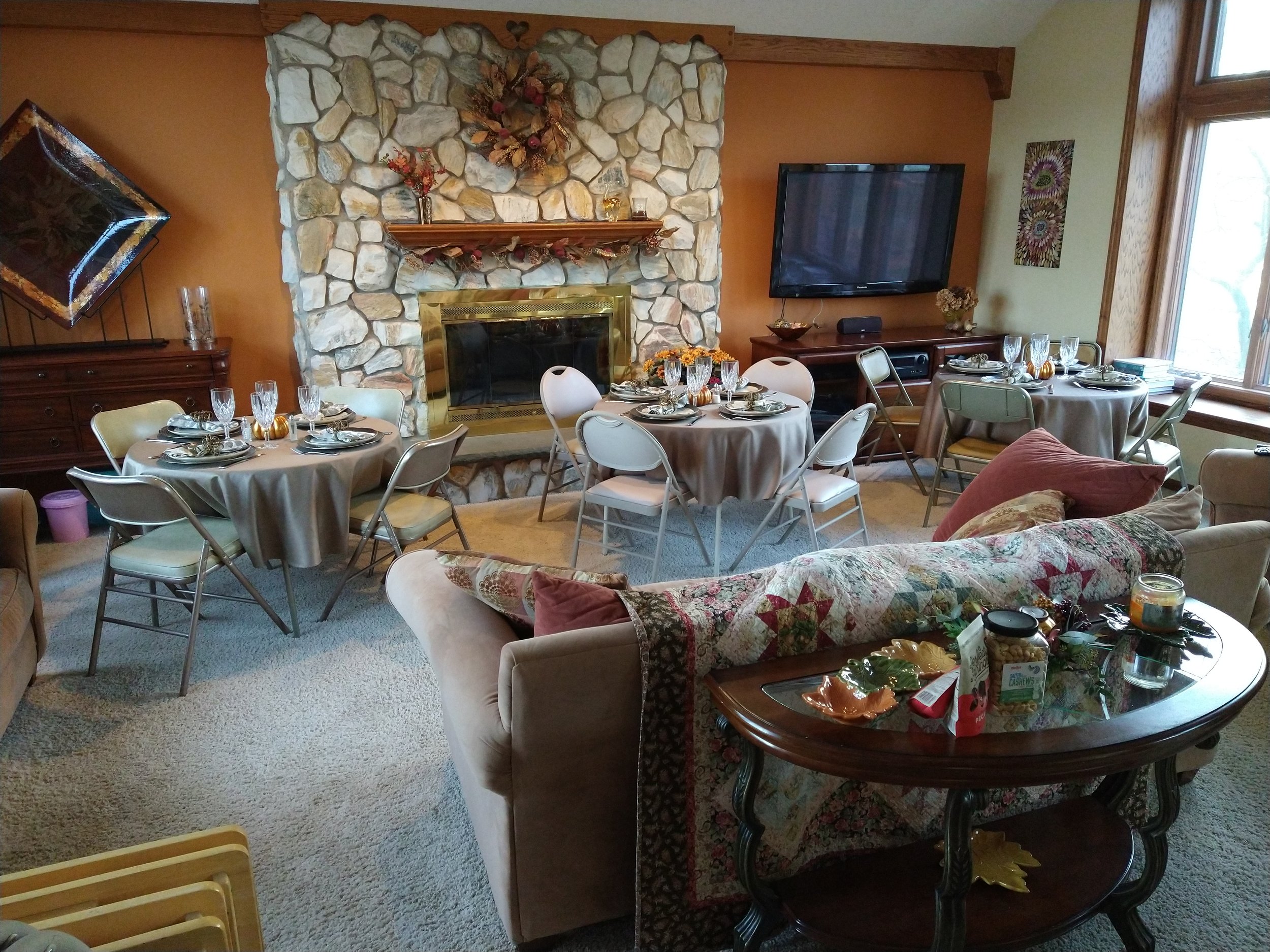 Living room with stone fireplace decorated for holiday, dining tables with place settings, a wall-mounted TV, and large windows with natural light.