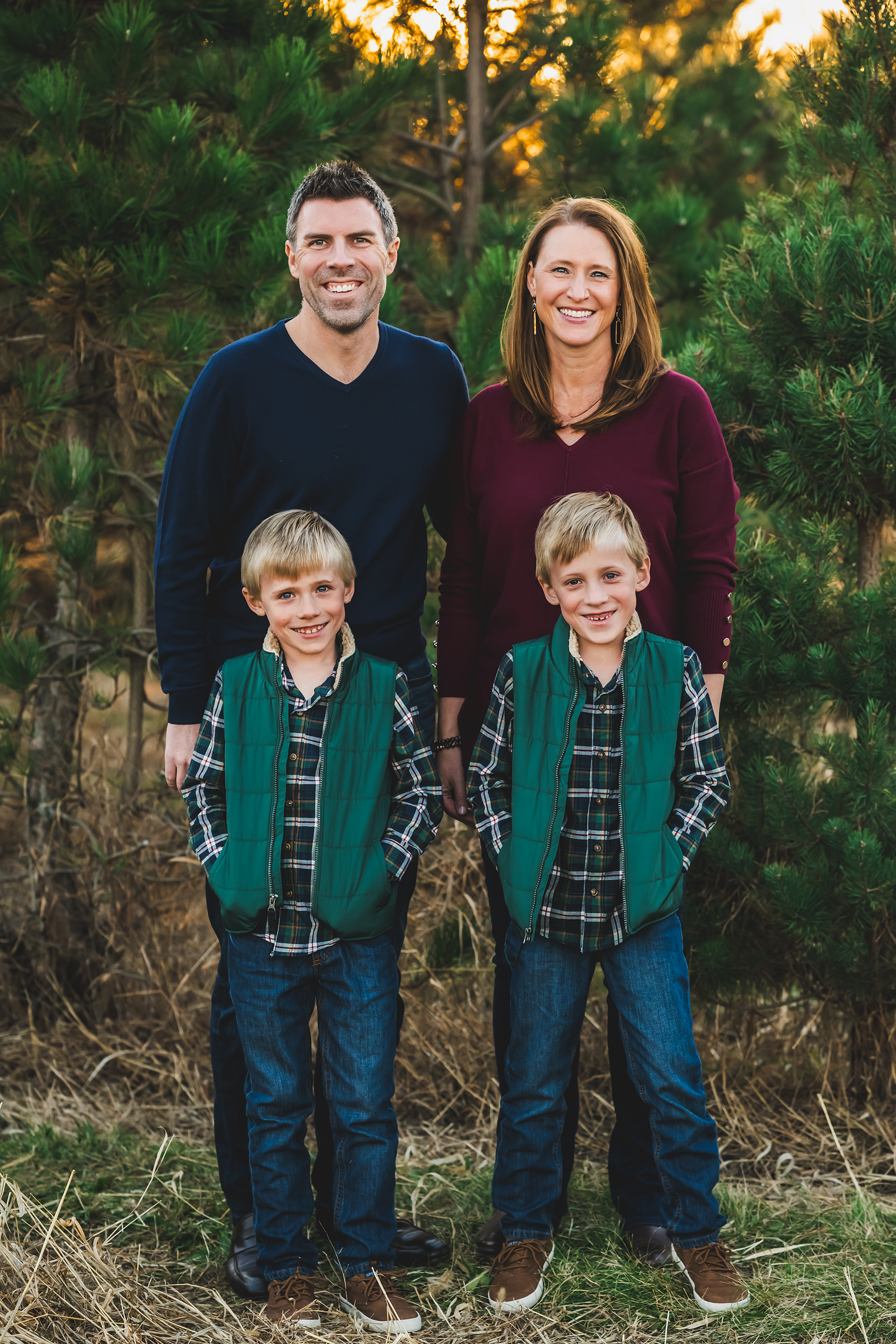 A family of four smiling outdoors in front of trees at sunset. The father and mother stand behind two young boys, all dressed casually in jackets and jeans.