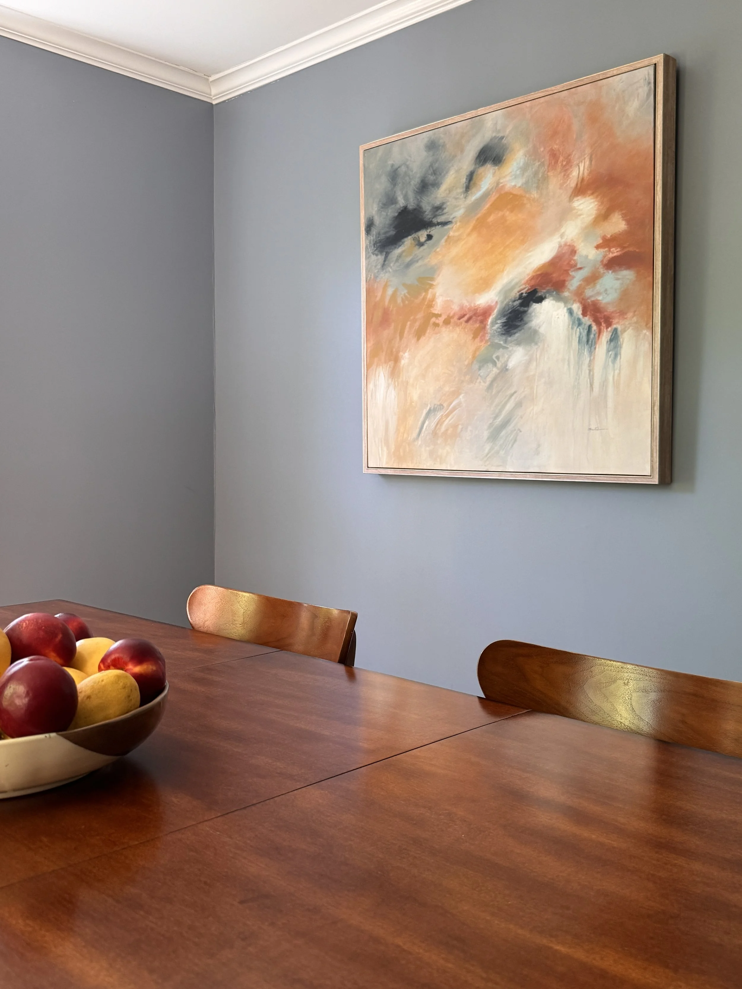 A wooden dining table with a bowl of apples and peaches, surrounded by wooden chairs, in a room with gray walls and a colorful abstract painting hanging on the wall.
