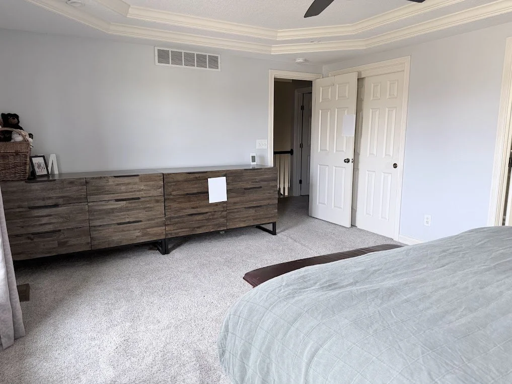 A bedroom with light gray walls, a beige carpet, a wooden dresser, an open closet door, and part of a bed with a gray quilt.