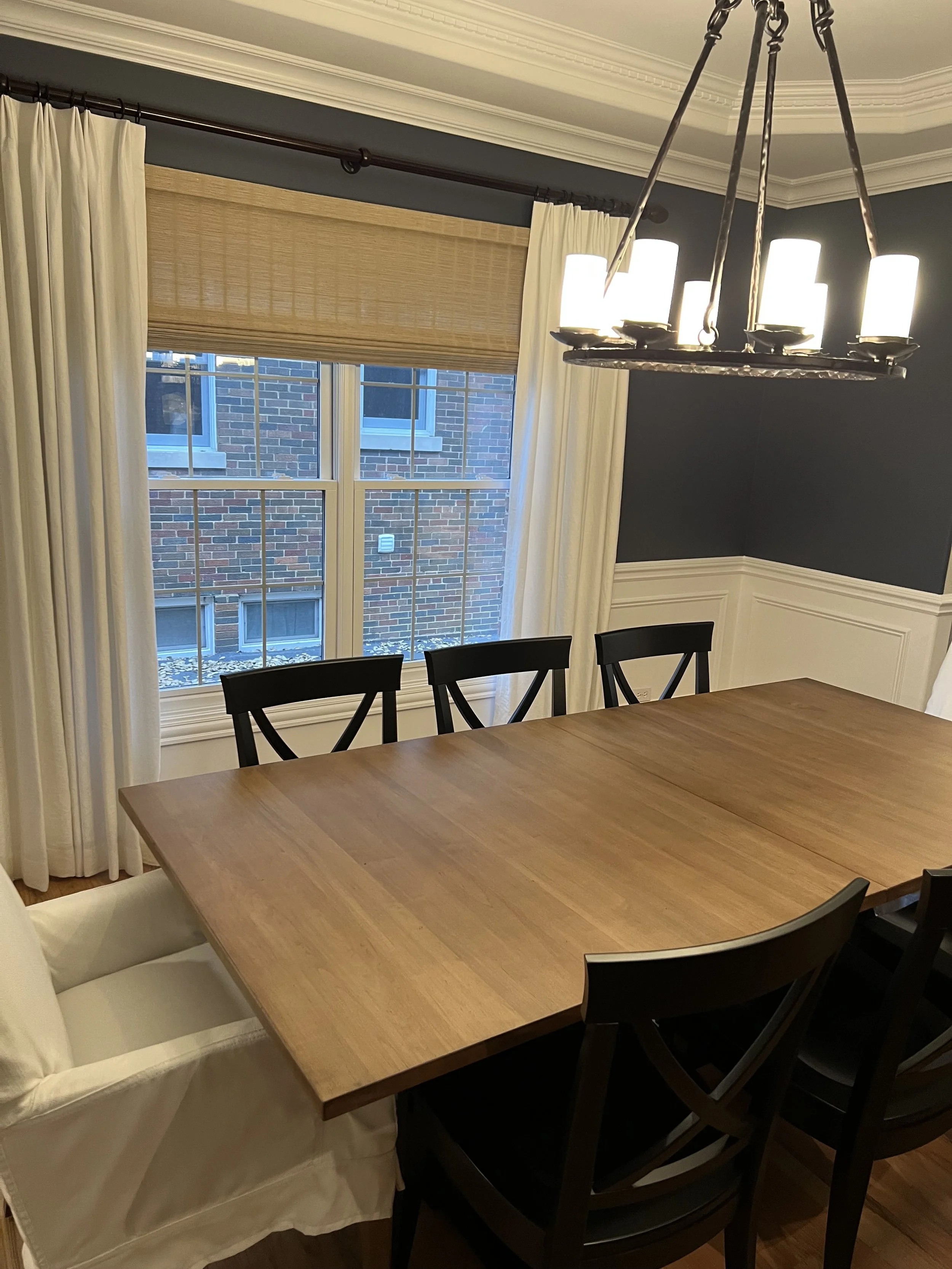 A wooden dining table surrounded by six black chairs, with a white cushioned armchair on one side, in a dining room with a window, curtains, and a chandelier with multiple lights.