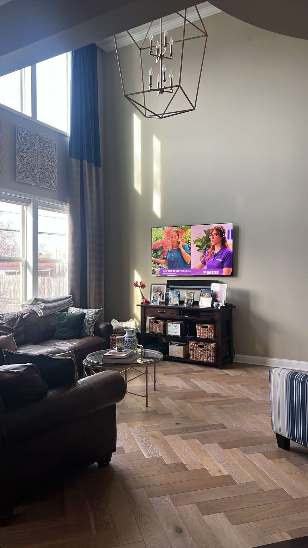 Living room with leather sofas, a glass coffee table, a TV on a wooden console, and large windows with blue and gray curtains. A chandelier hangs from the ceiling.