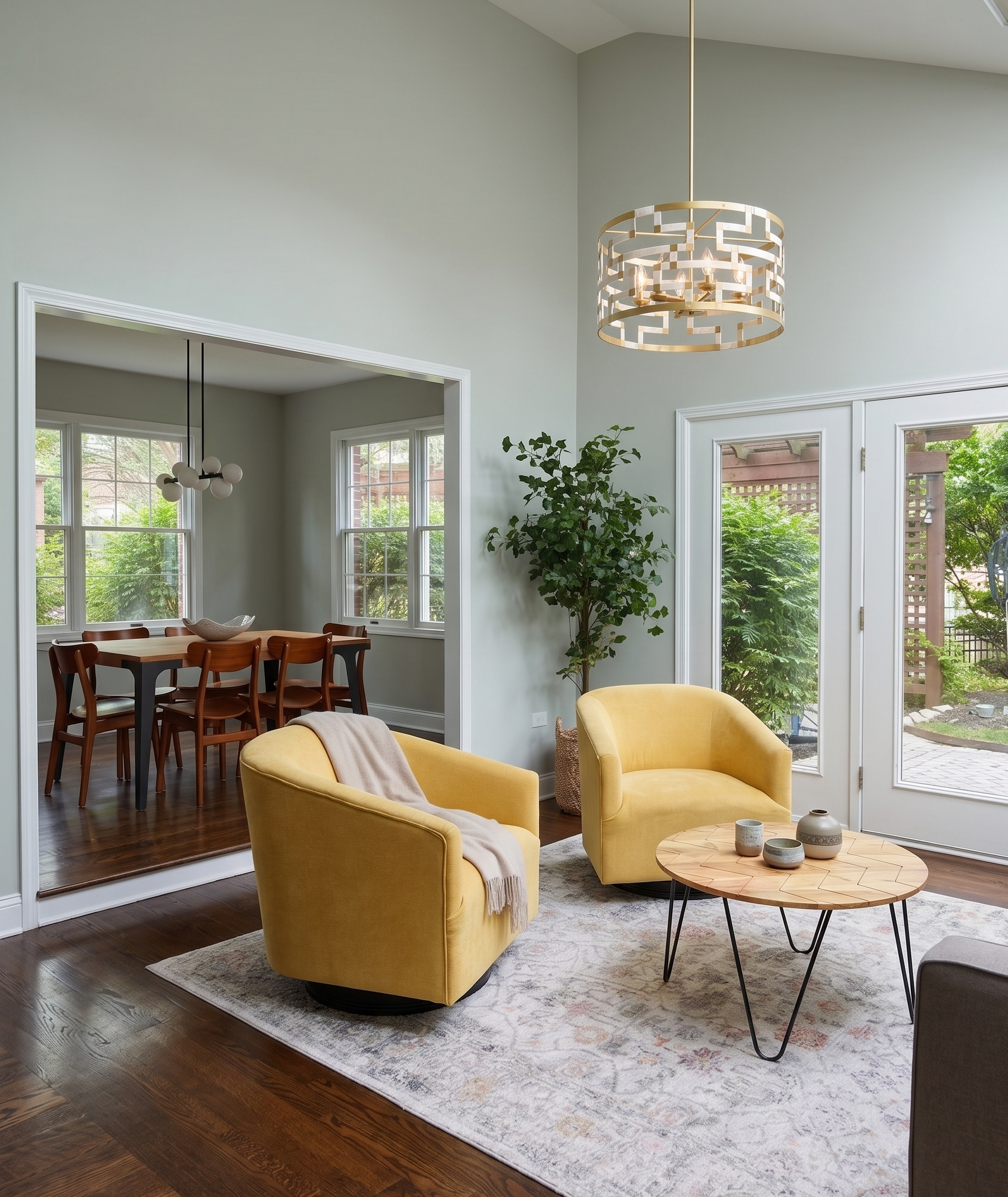 A bright, modern living room with yellow armchairs, a round wooden coffee table with decorative vases, and large windows with a view of greenery outside. There is an adjacent dining area with a wooden table and chairs, a contemporary chandelier, and a potted plant near the window.