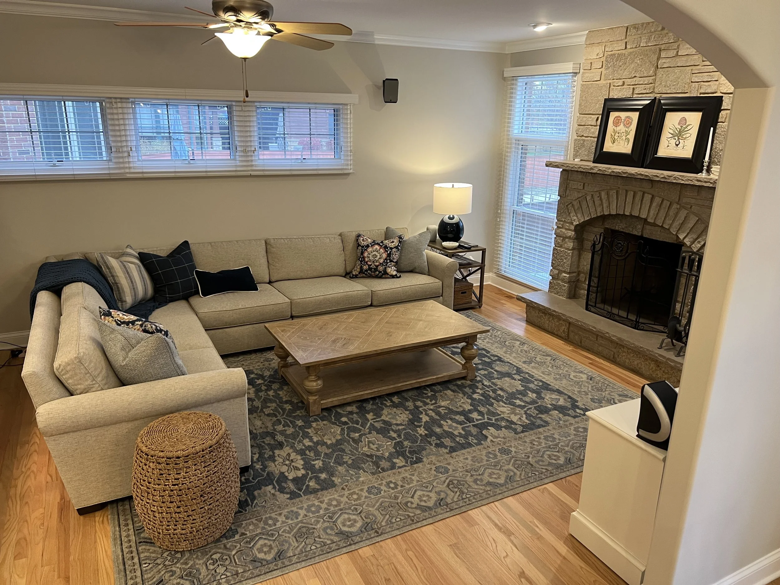 Living room with beige sectional sofa, black and floral throw pillows, wooden coffee table, area rug, stone fireplace with framed floral botanical prints, lamp, ceiling fan, and large windows with blinds.