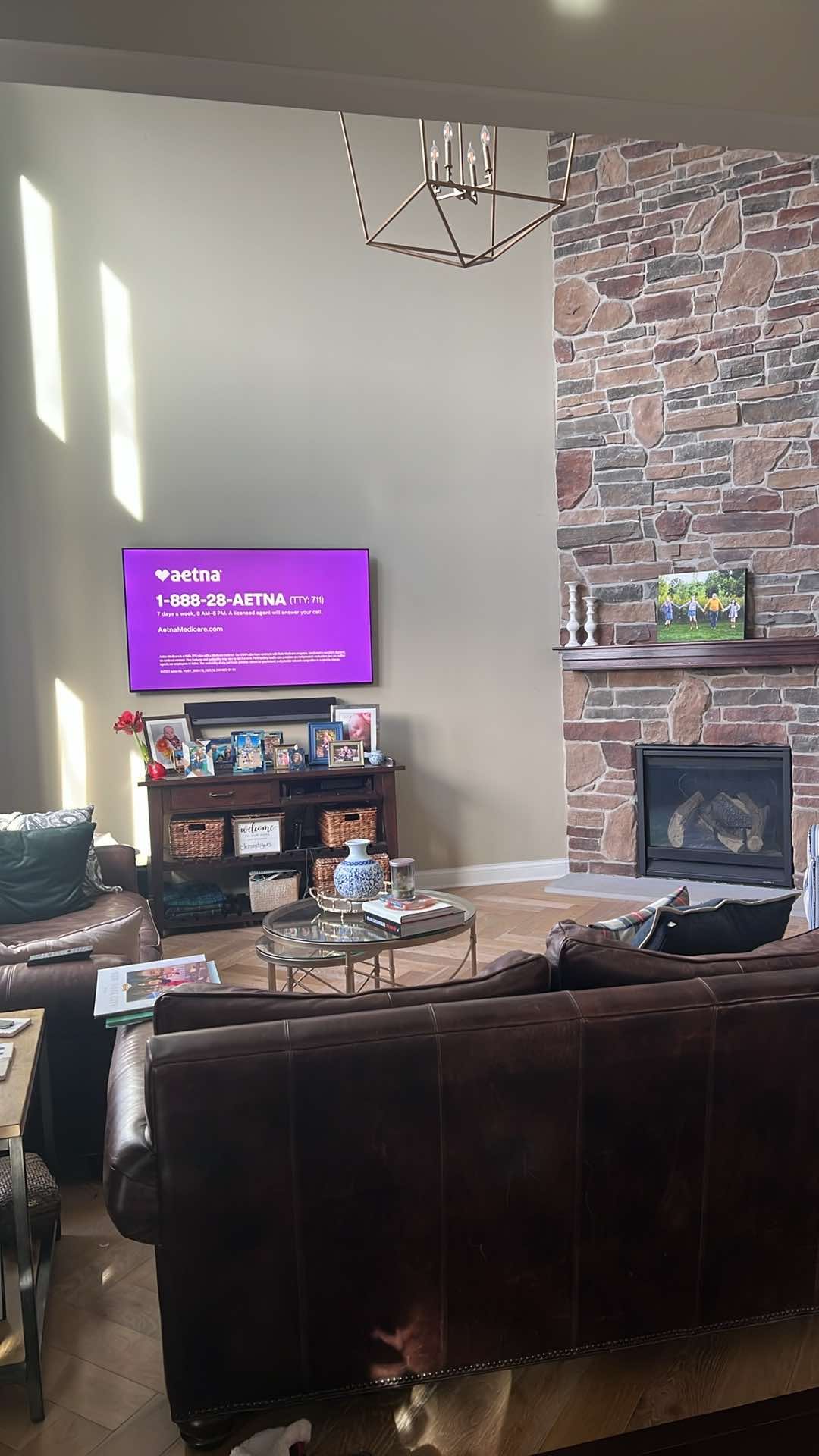 Living room with a leather couch, a wooden entertainment center with family photos, a television displaying an Aetna advertisement, a stone fireplace, and a side table with decorative items, illuminated by sunlight through windows.