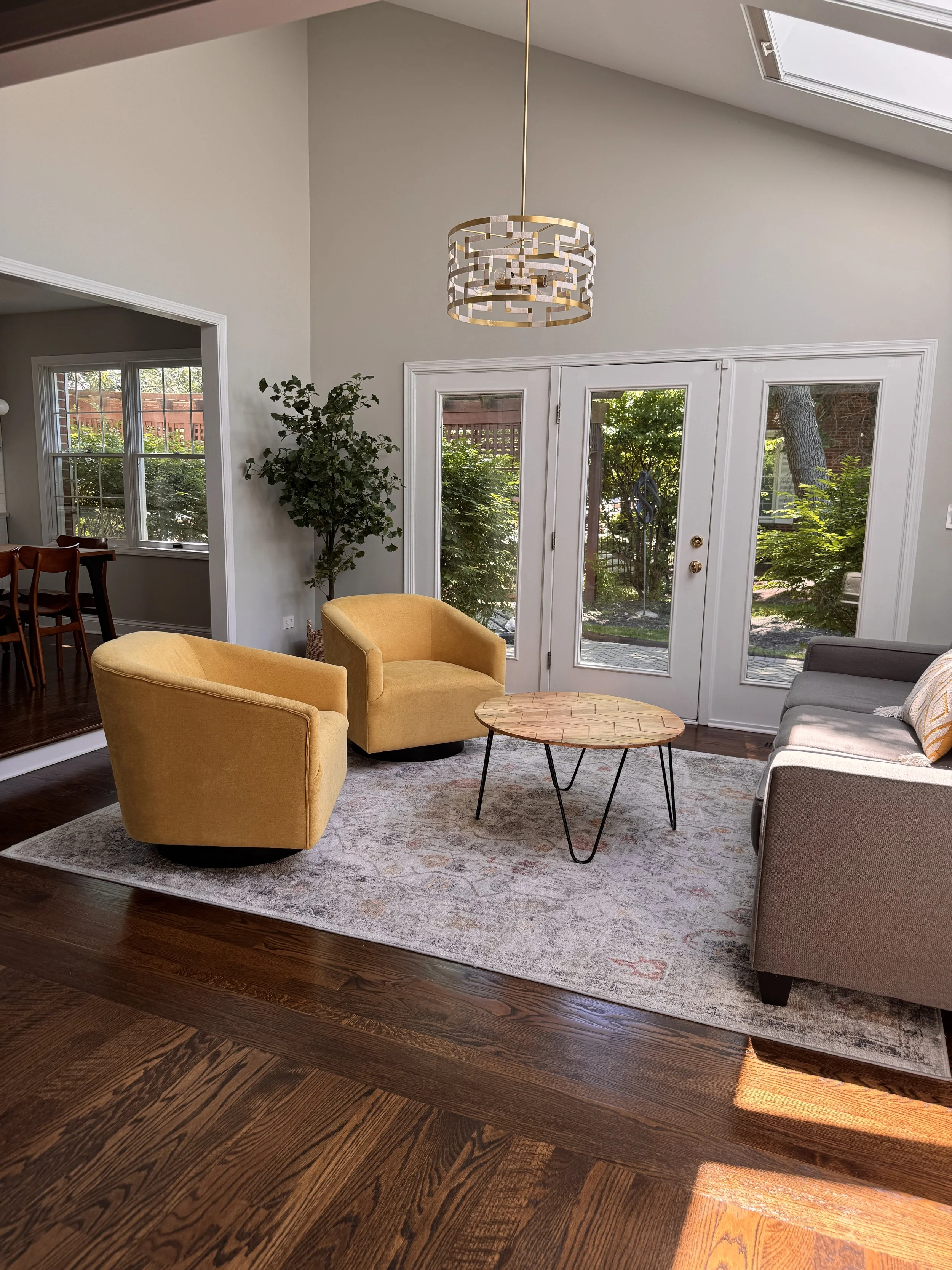 Living room with two yellow armchairs, a round wooden coffee table, a gray sofa with pillows, a large area rug, a potted plant, and glass doors leading outside, with sunlight coming in.