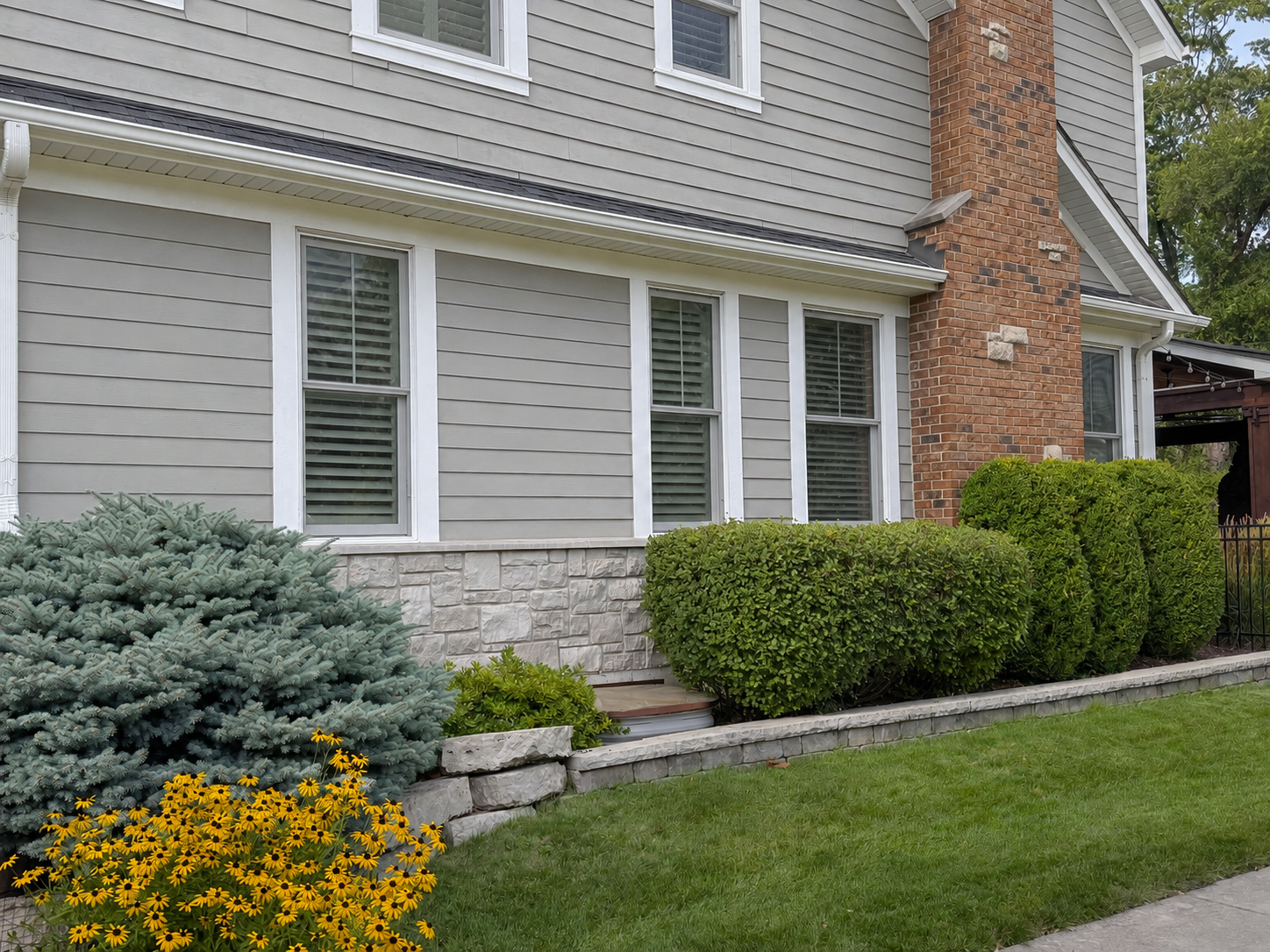 Exterior view of a house with gray siding, white trim, and a brick chimney. There are four windows with white shutters and landscaped bushes and flowers in front, including yellow flowers and a blue-green conifer.