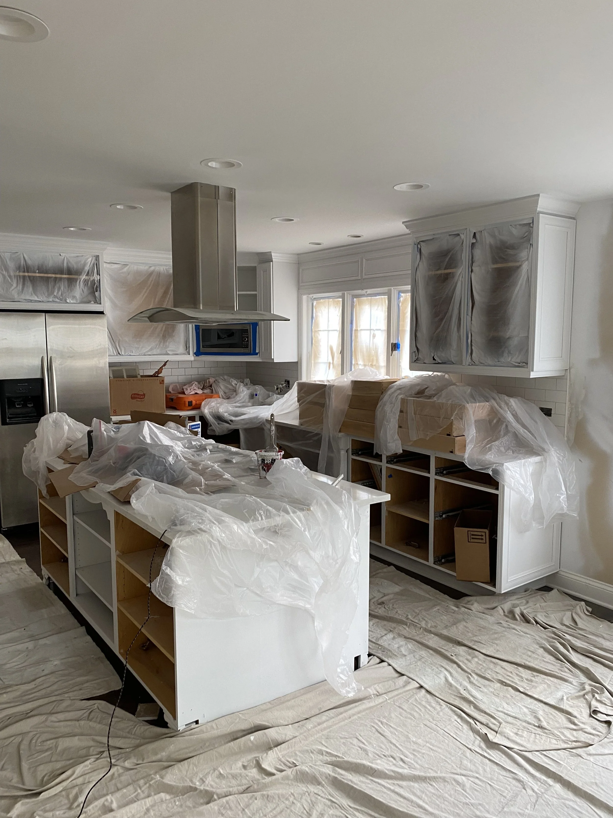 A kitchen under renovation with cabinets covered in plastic, a kitchen island surrounded by plastic, and construction tools and materials scattered on the floor and countertops.