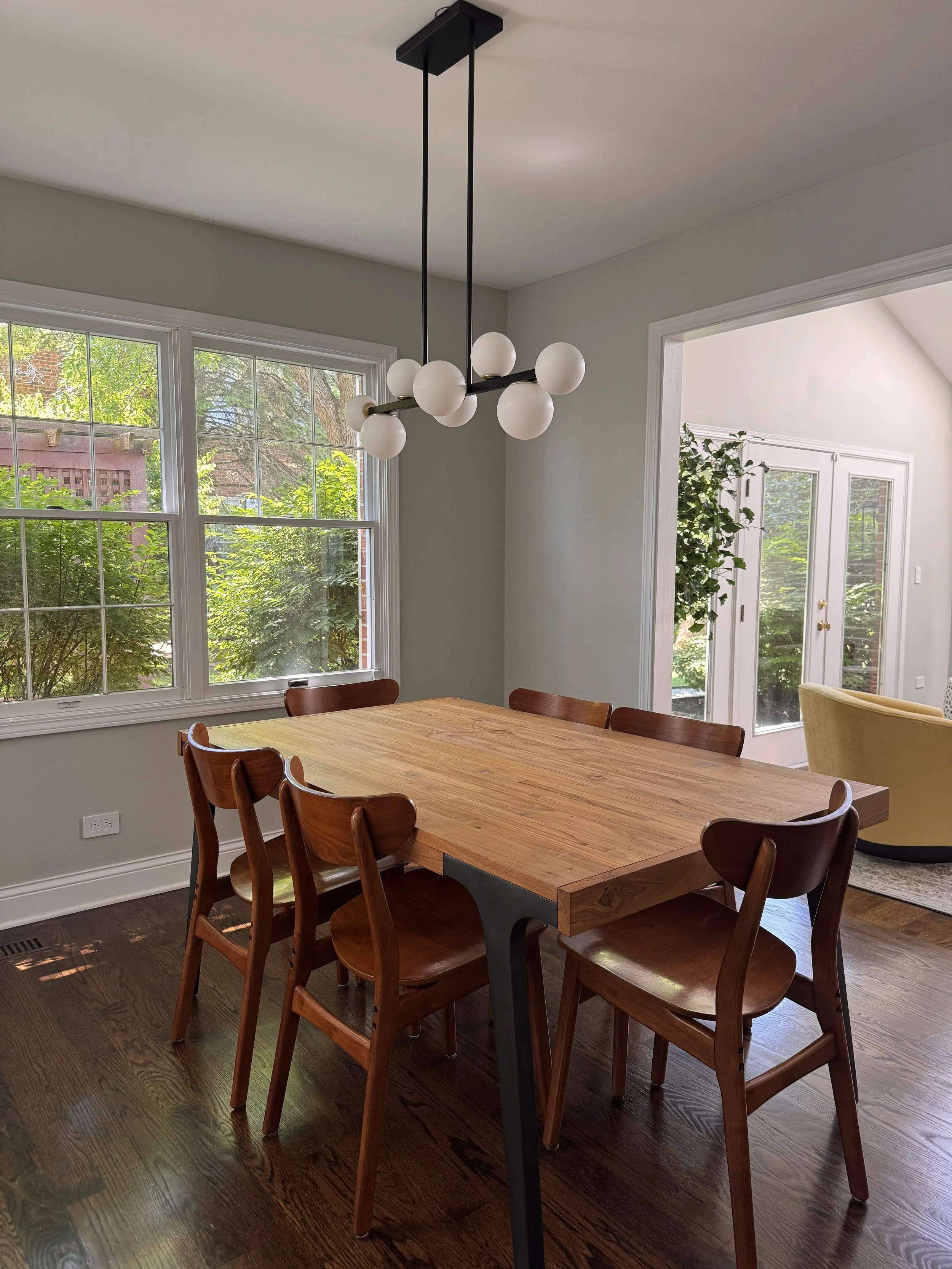 A dining room with a wooden table, eight wooden chairs, large windows showing greenery outside, and a modern chandelier with white globe lights hanging from the ceiling.