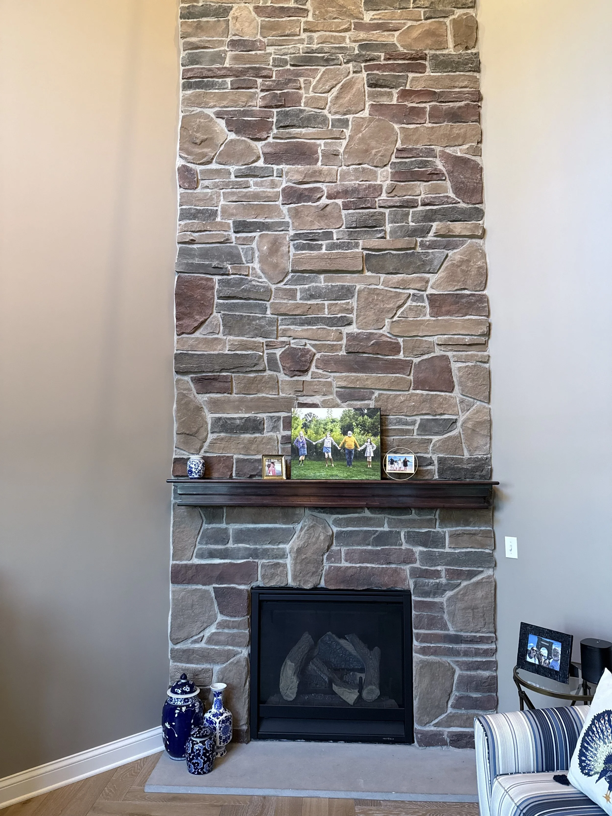 A stone fireplace with a wood mantel, decorated with three blue and white vases and framed photos. To the right, a striped armchair with pillows, and to the right, a small table with framed photos and a speaker.