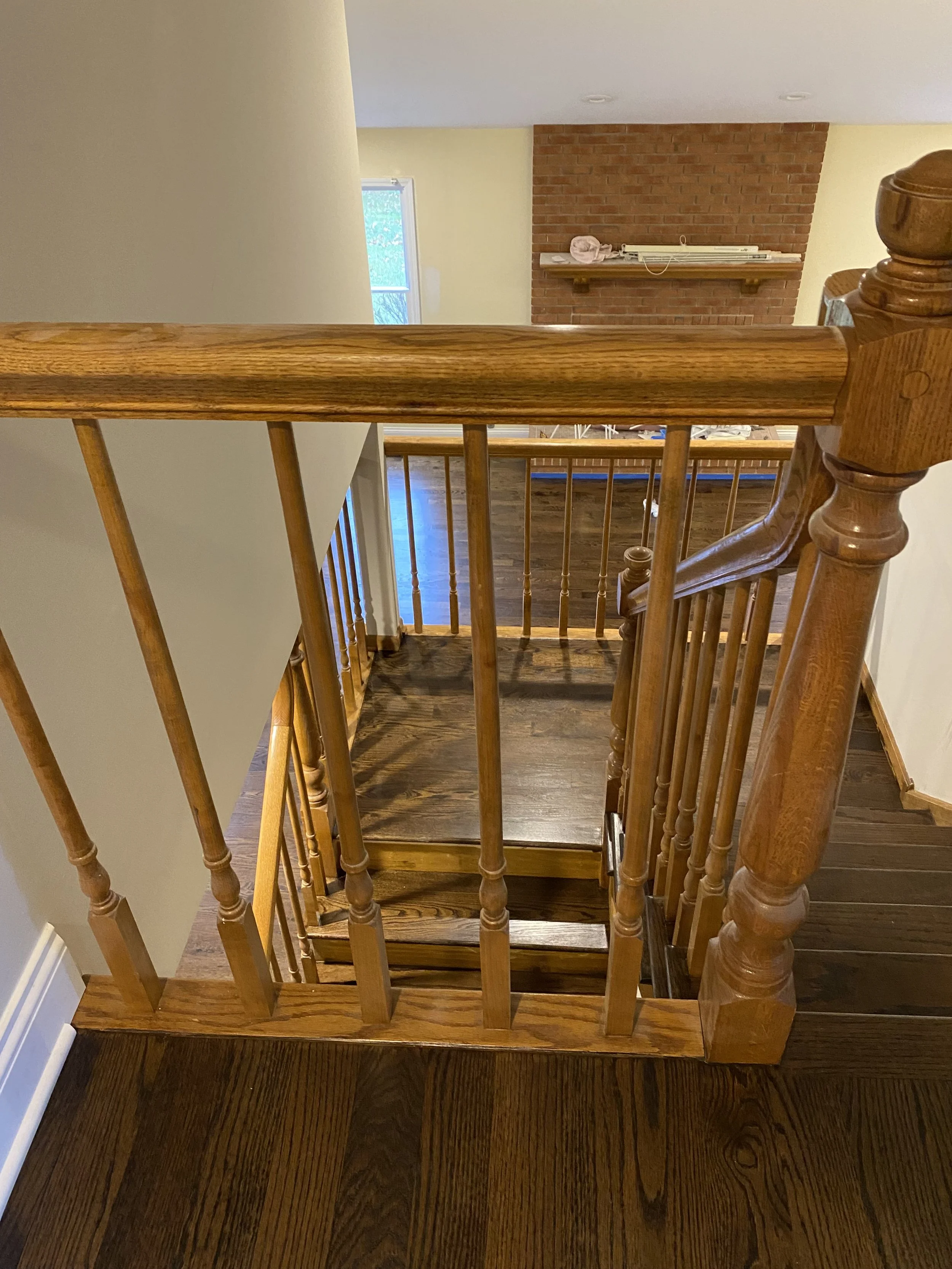 View from the top of a wooden staircase looking down to the lower level with hardwood flooring, brick fireplace, and a window with natural light.