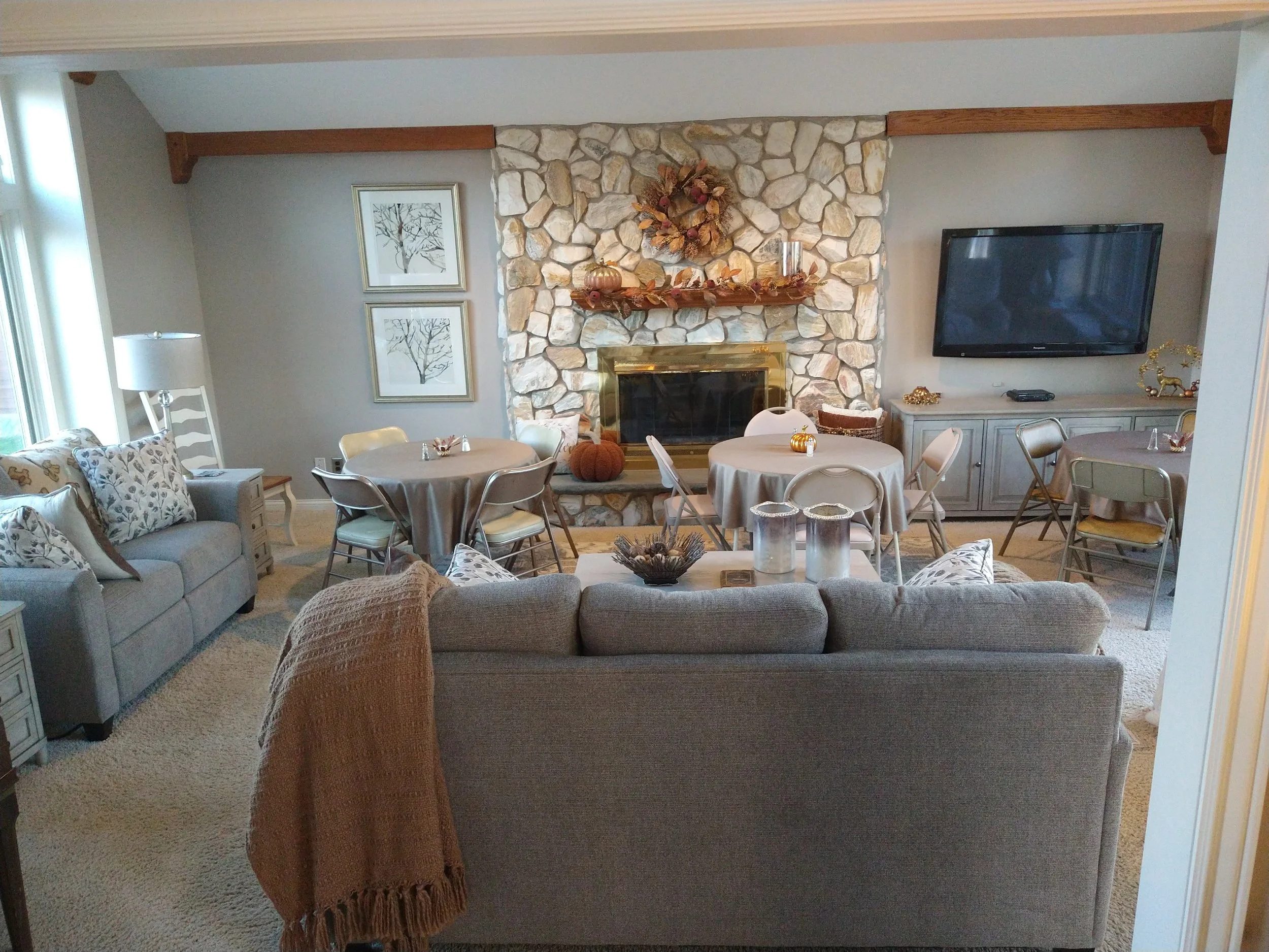 Living room with a stone fireplace, decorated for fall with pumpkins and autumn leaves, framed artwork of trees on the wall, and a large flat-screen TV on a cabinet.