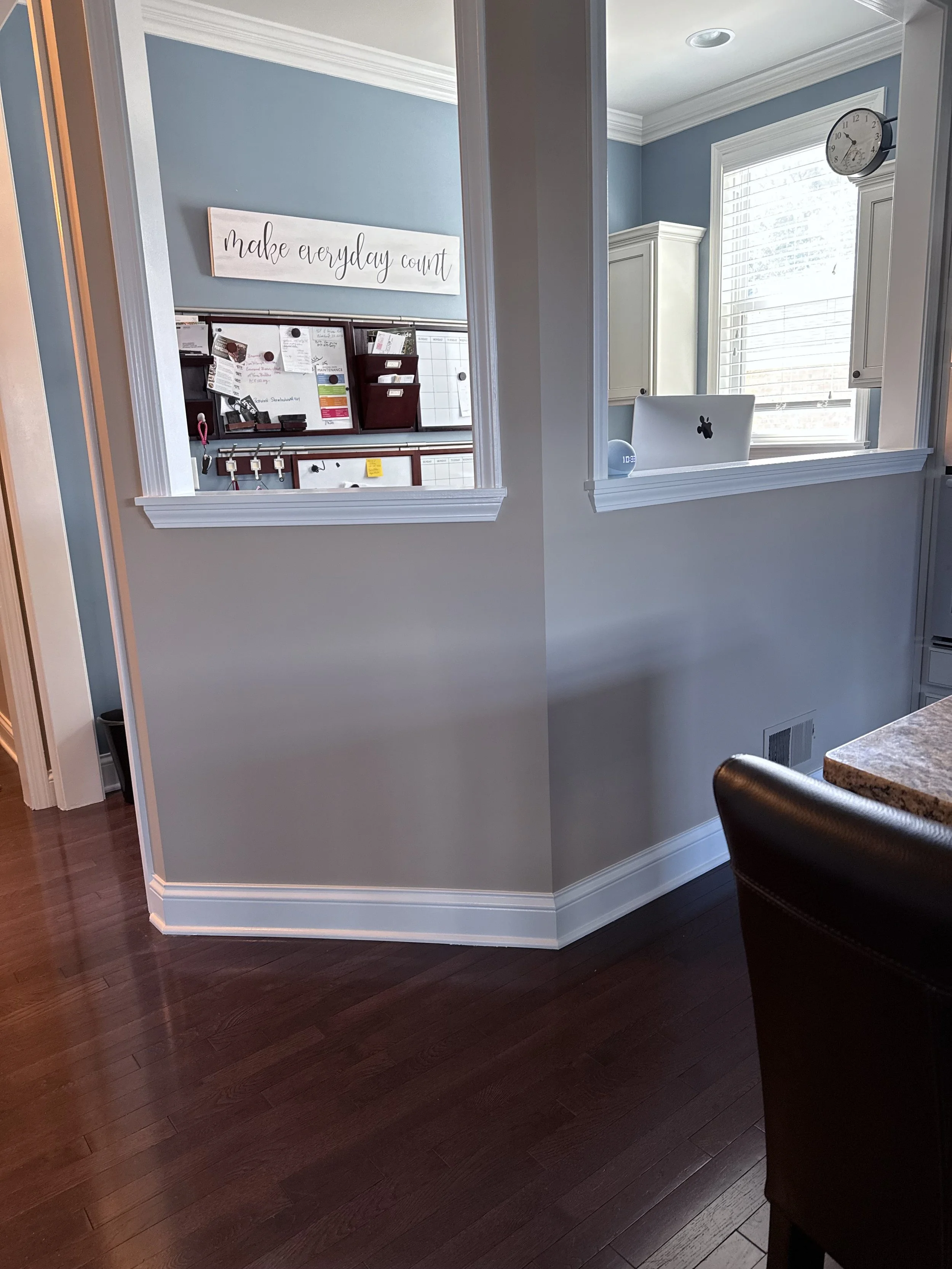 View of a home interior wall with a window and a clock, with a kitchen counter and chair in the foreground, and a visible office or workspace through a doorway, including a bulletin board, sign with quote, and a MacBook.