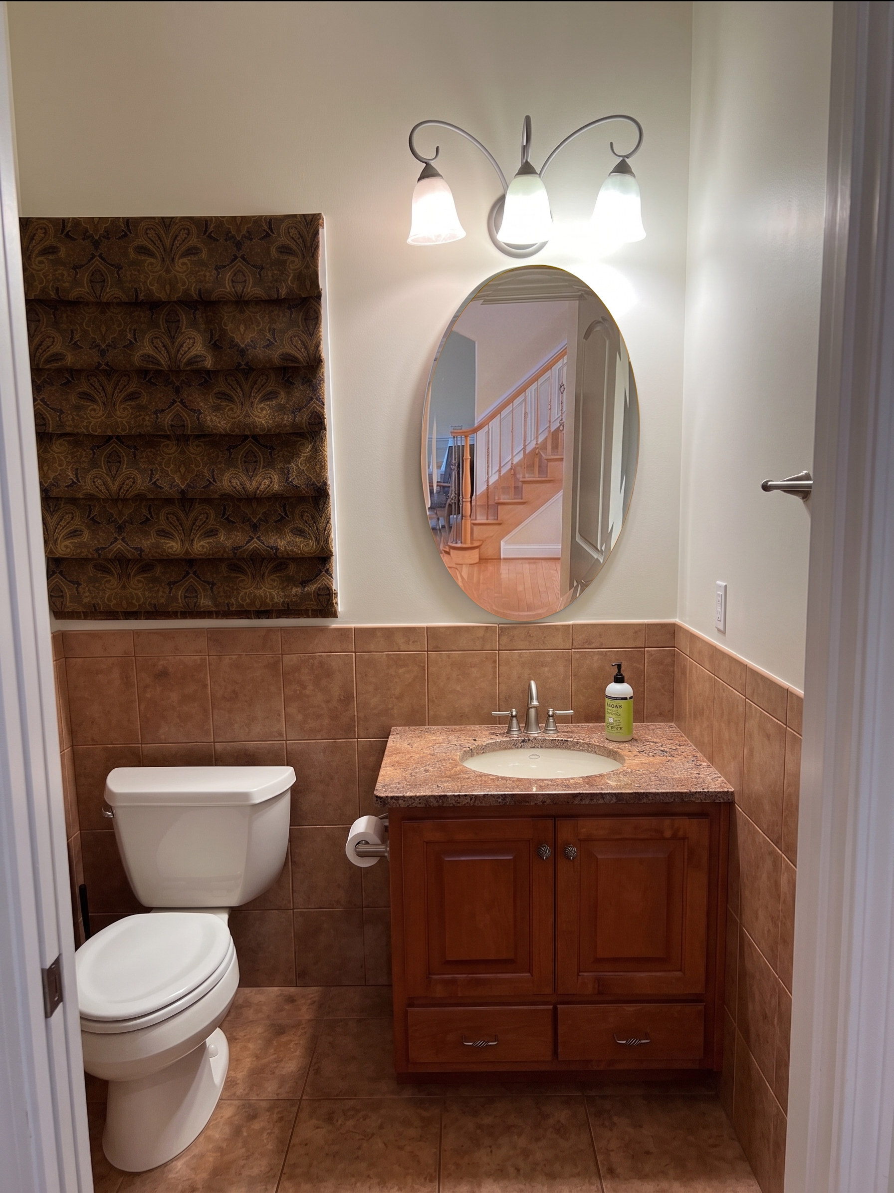 Bathroom with a toilet, sink with wooden cabinet, oval mirror, wall-mounted light fixture with three lamps, and brown tiled walls and floors.