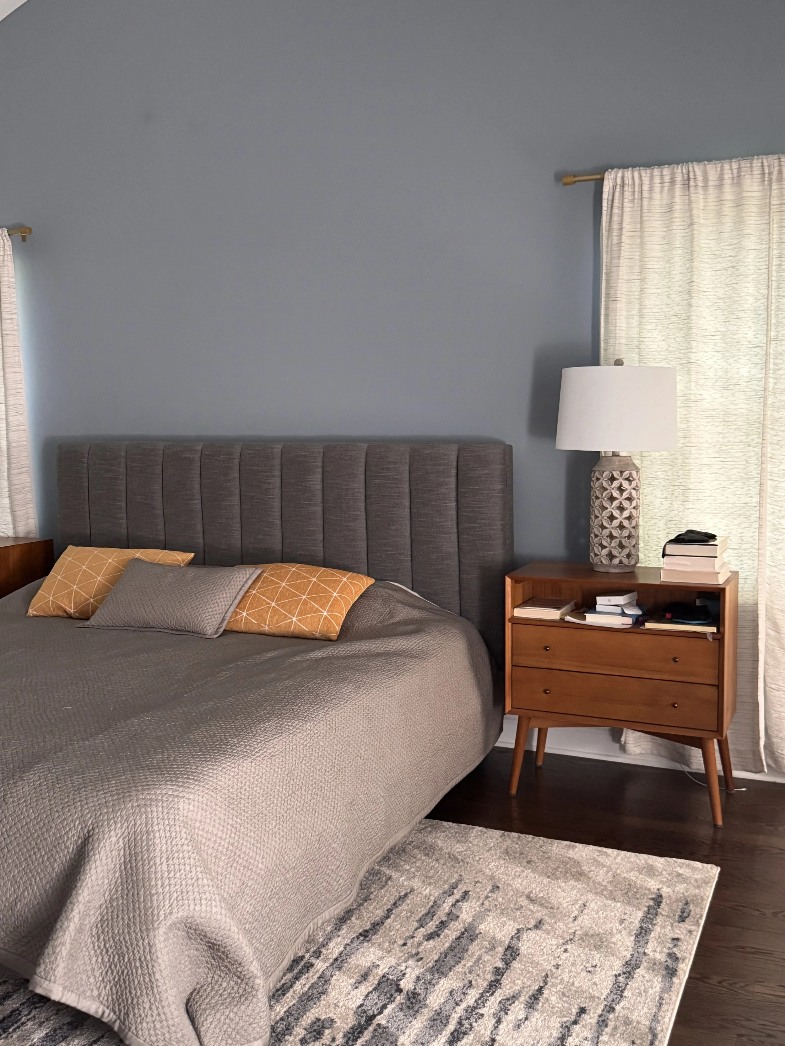 A cozy bedroom featuring a large bed with a gray headboard, gray bedspread, and multiple pillows in shades of orange, beige, and gray. Next to the bed, there is a wooden nightstand with a decorative table lamp, books, and a phone. The room has a large window with cream-colored curtains, a textured area rug with blue, gray, and cream tones, and dark hardwood floors.