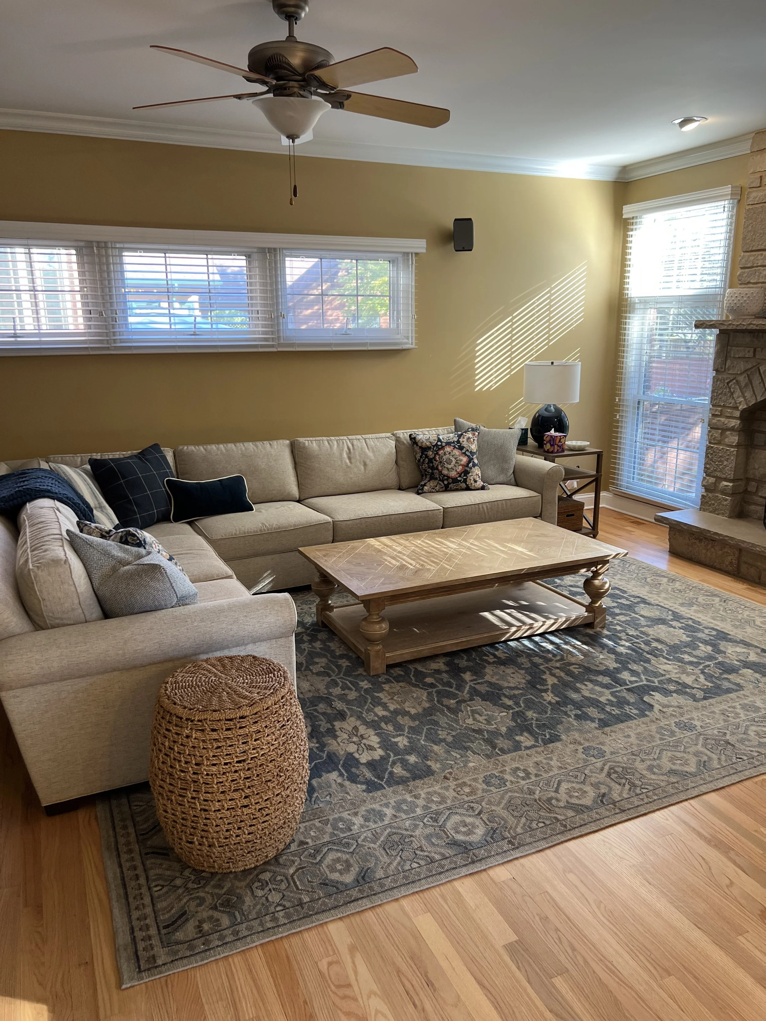 A cozy living room with beige L-shaped sofa decorated with patterned and solid colored pillows. A wooden coffee table sits in front. An ornate area rug covers part of the hardwood floor. A woven ottoman is in the foreground. A TV lamp and a tissue box are on a side table next to the sofa. There are large windows with blinds allowing sunlight to cast shadows on the wall. A ceiling fan is above, and a stone fireplace is on the right.
