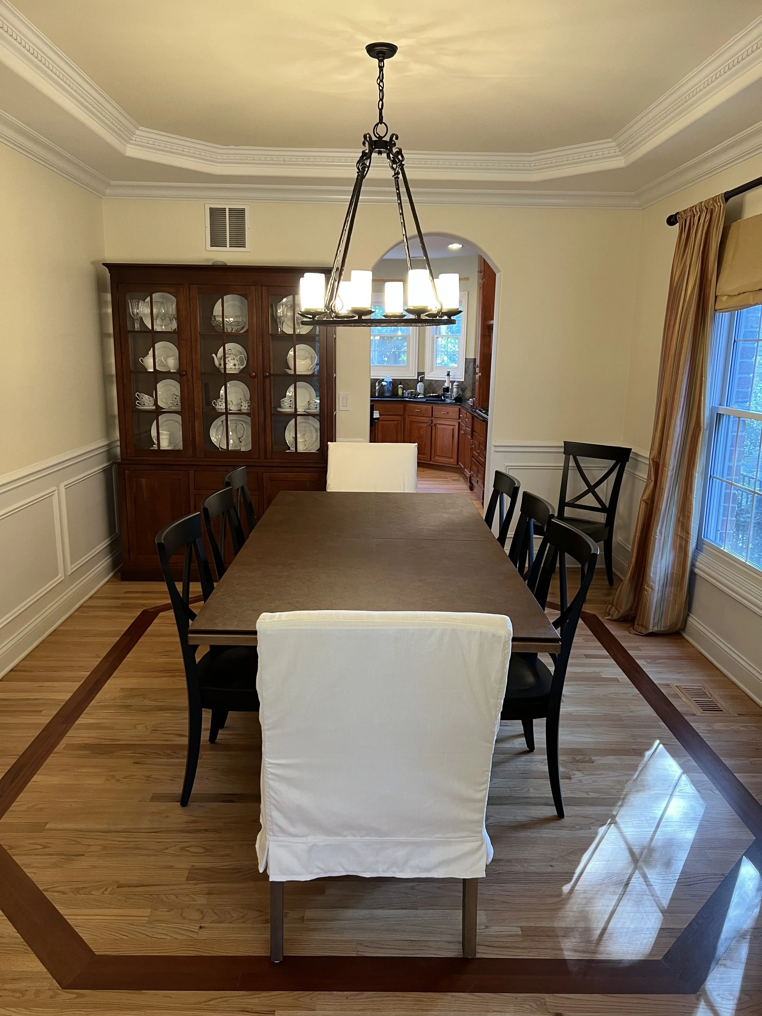 A dining room with a large dining table, six black chairs, a china cabinet filled with white dishes, a chandelier hanging from the ceiling, and a window with curtains.