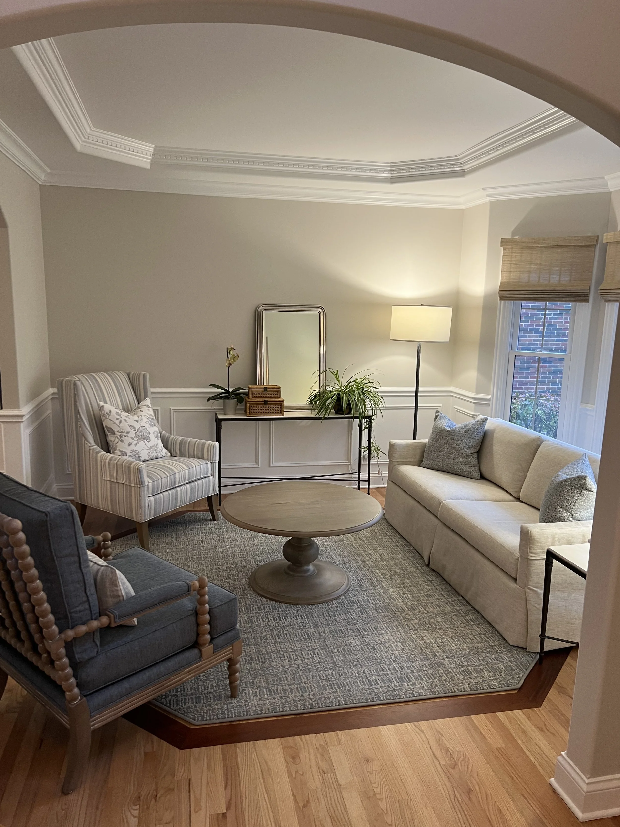 A living room with a beige sofa, patterned armchair, round coffee table, side table with greenery and mirror, window with wooden blinds, and a floor lamp near the window. The room has light-colored walls, crown molding, and hardwood flooring.