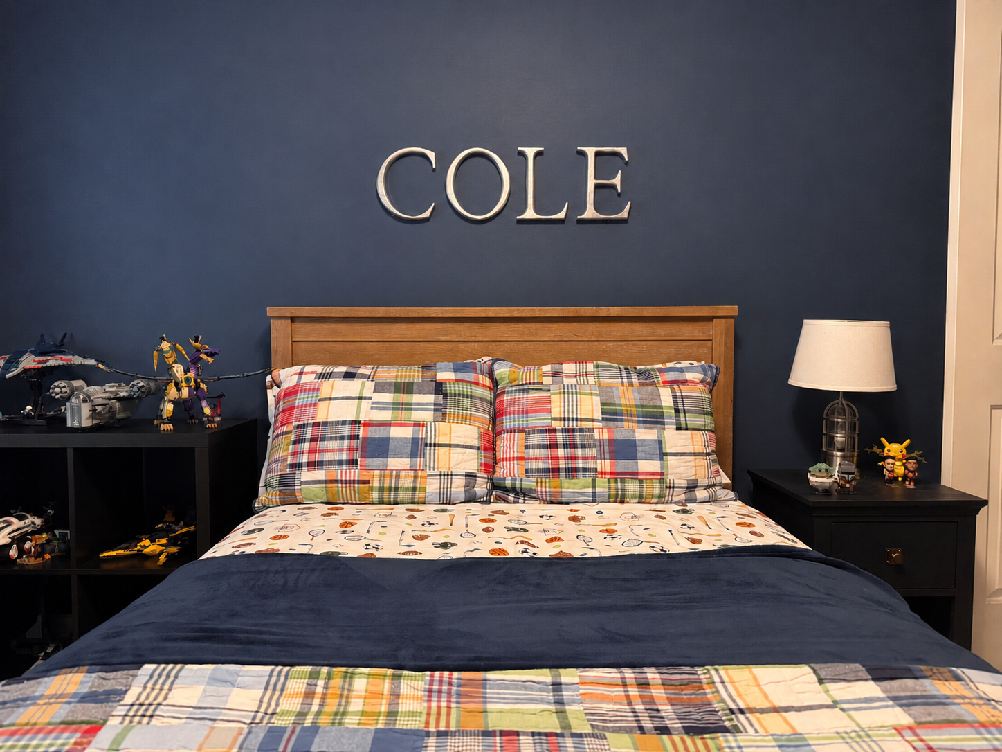 Bedroom with a wooden bed, colorful plaid pillows, Mario-themed bedding, blue wall with the name 'COLE' in large letters, black nightstand with toys and a lamp, and a black shelf with toys including Transformers and LEGO figures.