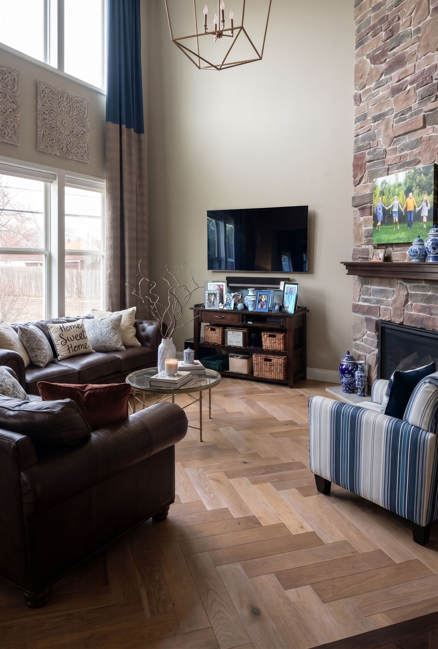 Living room with large window, high ceiling, chandelier, brick fireplace, sofa with decorative pillows, striped armchair, coffee table, and TV, filled with natural light.