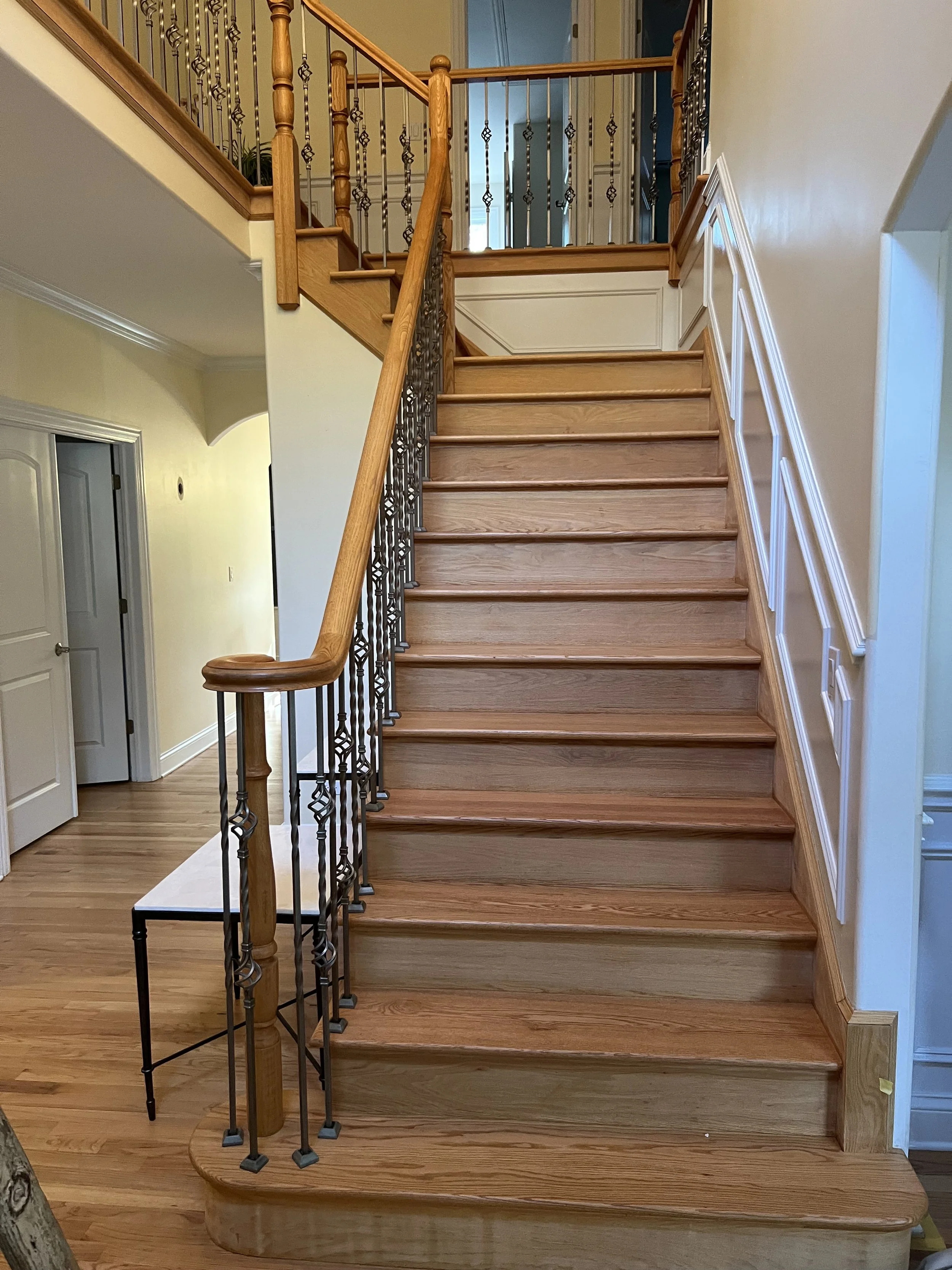 Wooden staircase with carpeted steps, dark wrought iron and wooden balusters, and a handrail leading to an upper floor. A small gray table is at the base of the staircase. A doorway to the left leads to another room, and the walls are painted light yellow with decorative white trim.