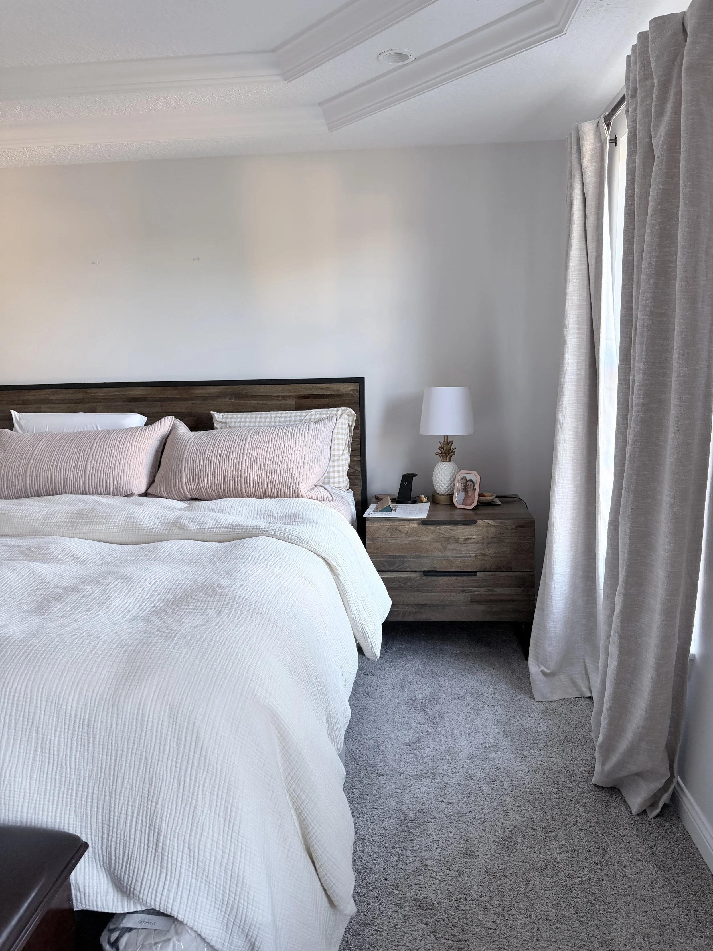 A bedroom with a bed that has a white quilt, pink pillows, and a dark headboard, next to a nightstand with a white lamp, framed photo, and phone, next to a window with closed curtains and a gray carpet.