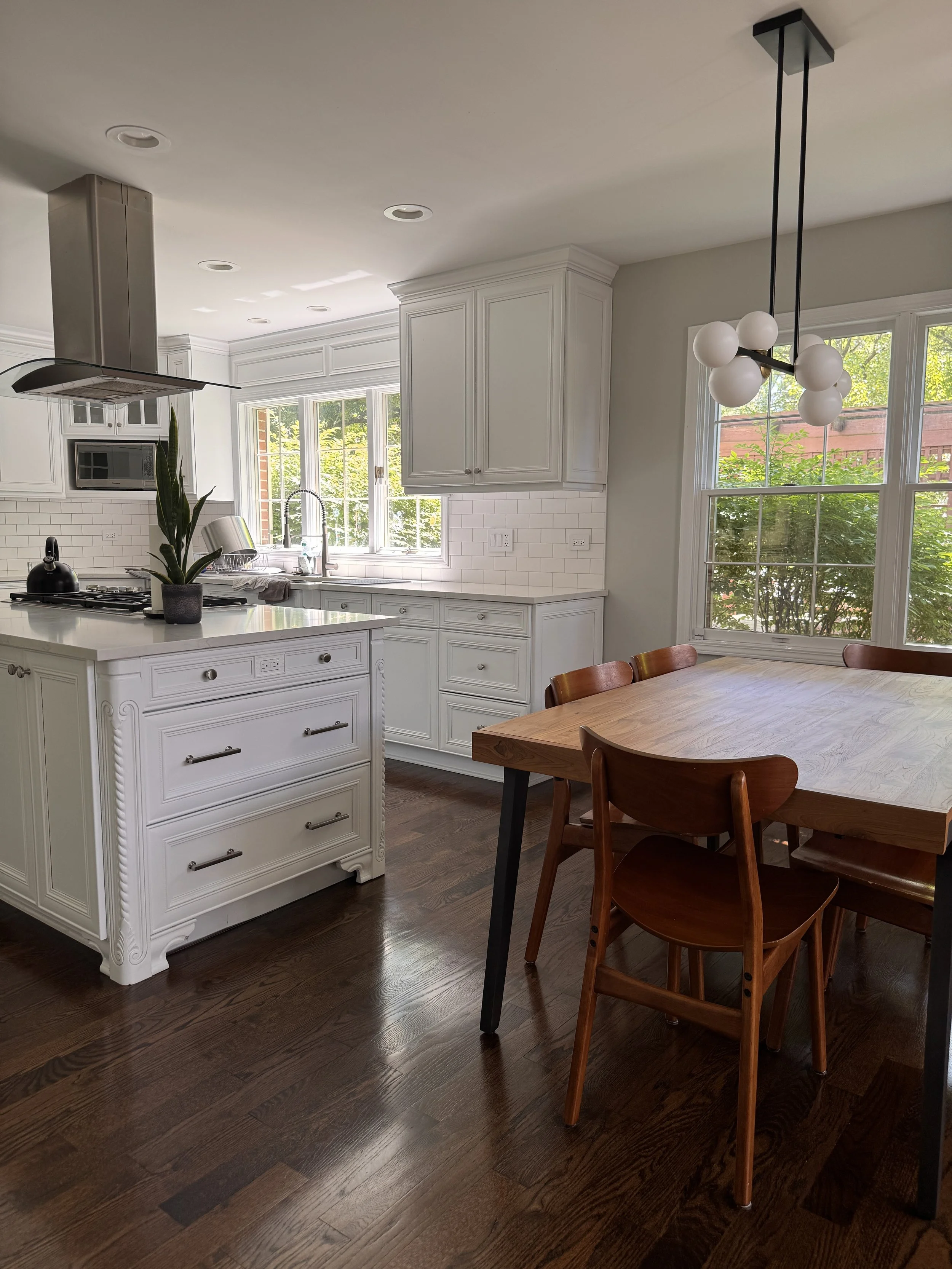 A bright kitchen with white cabinets, a white island with drawers, large windows, and a wooden dining table with matching chairs. There is a modern black and white chandelier hanging from the ceiling.
