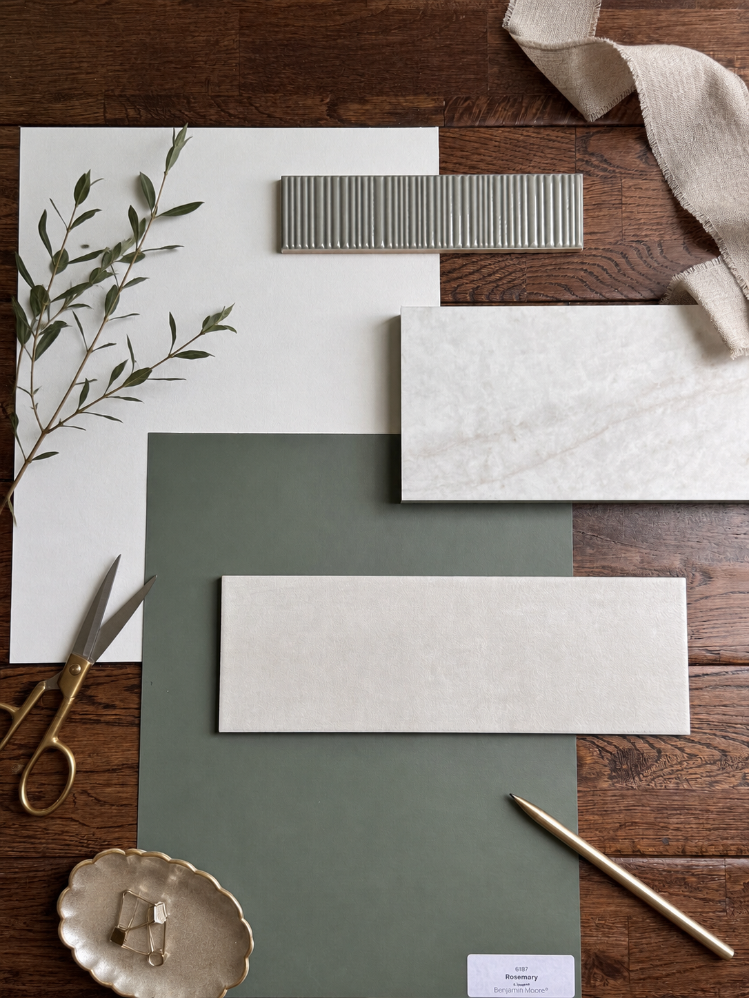 Interior design mood board with various tiles, green and white, a branch of rosemary, scissors, a gold pen, a beige cloth, and a small dish with clips, on a wooden surface.
