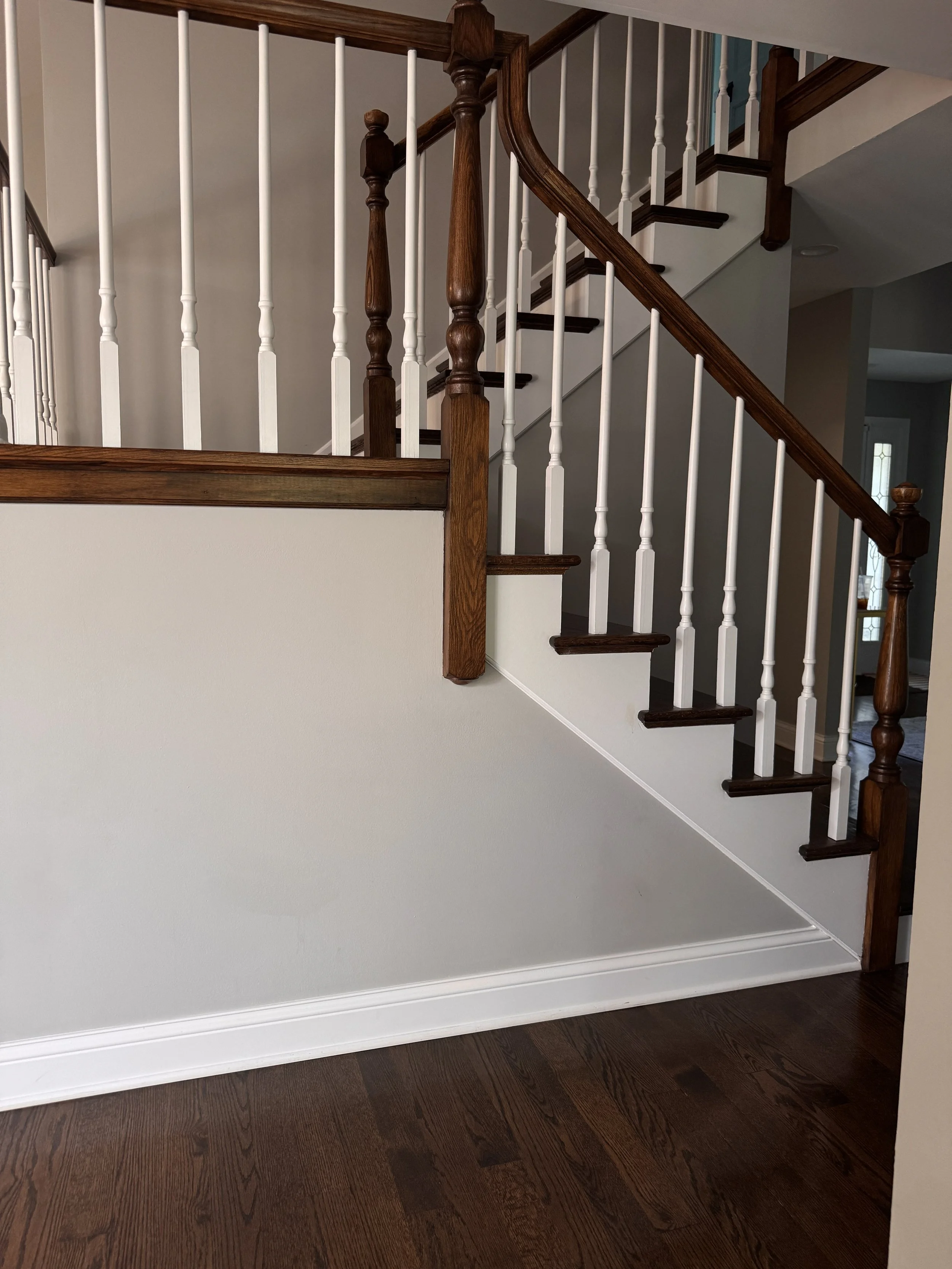 Interior view of a staircase with brown wooden steps, white spindles, and a brown wooden handrail, with a section of the main floor showing dark wood flooring.
